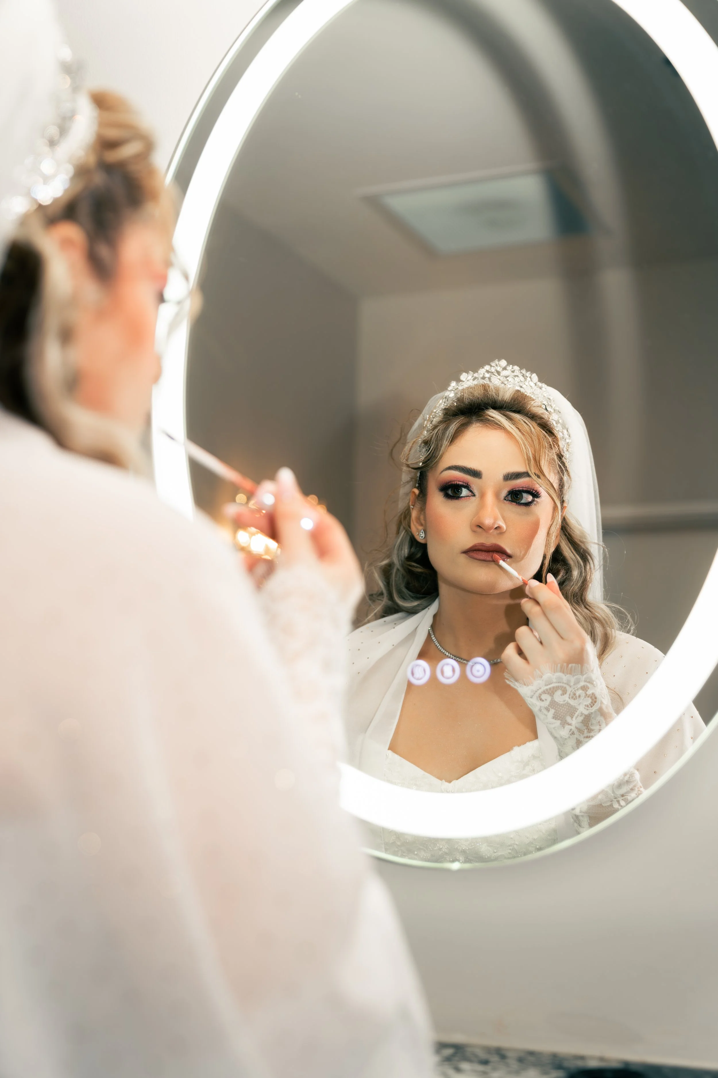 Bride in wedding dress and tiara applying lipstick while looking at her reflection in a mirror.