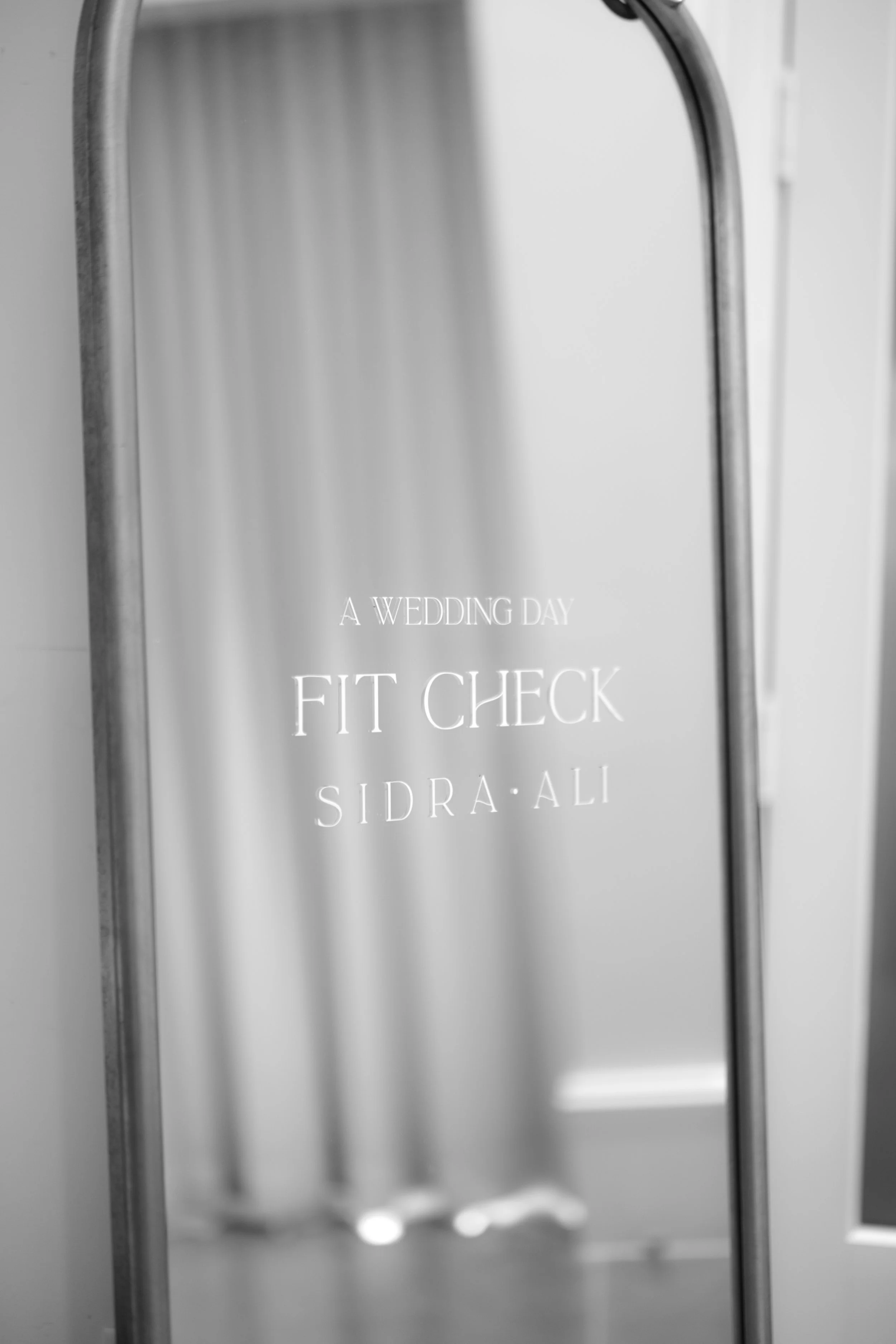 A sign with white text on a transparent background that reads: "A wedding day fit check Sidra Ali". The sign appears to be placed on a mirror or glass surface, with a metal frame visible on the sides.