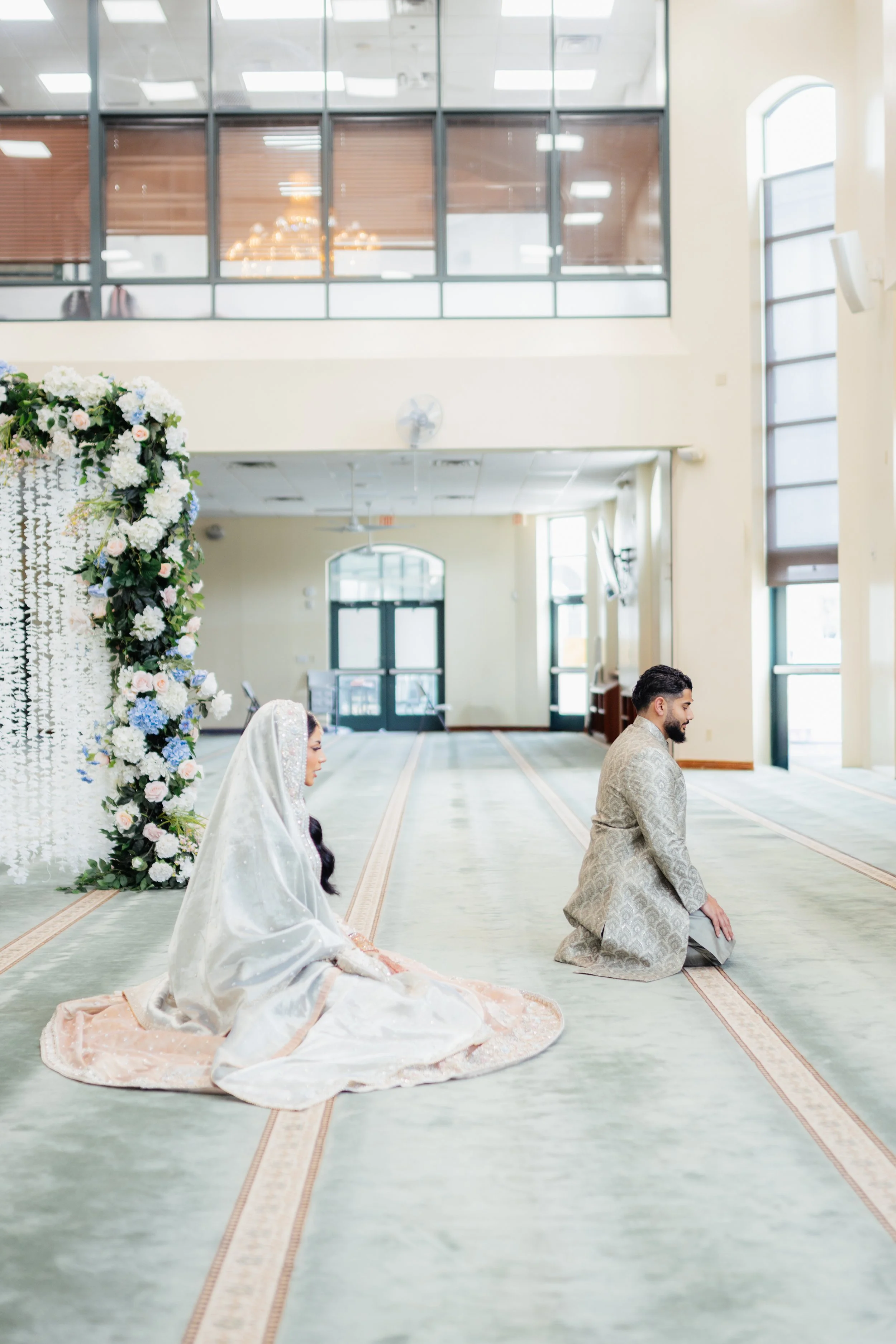 A bride and groom kneeling in prayer inside a spacious, well-lit room with large windows and a high ceiling, decorated with floral arrangements.