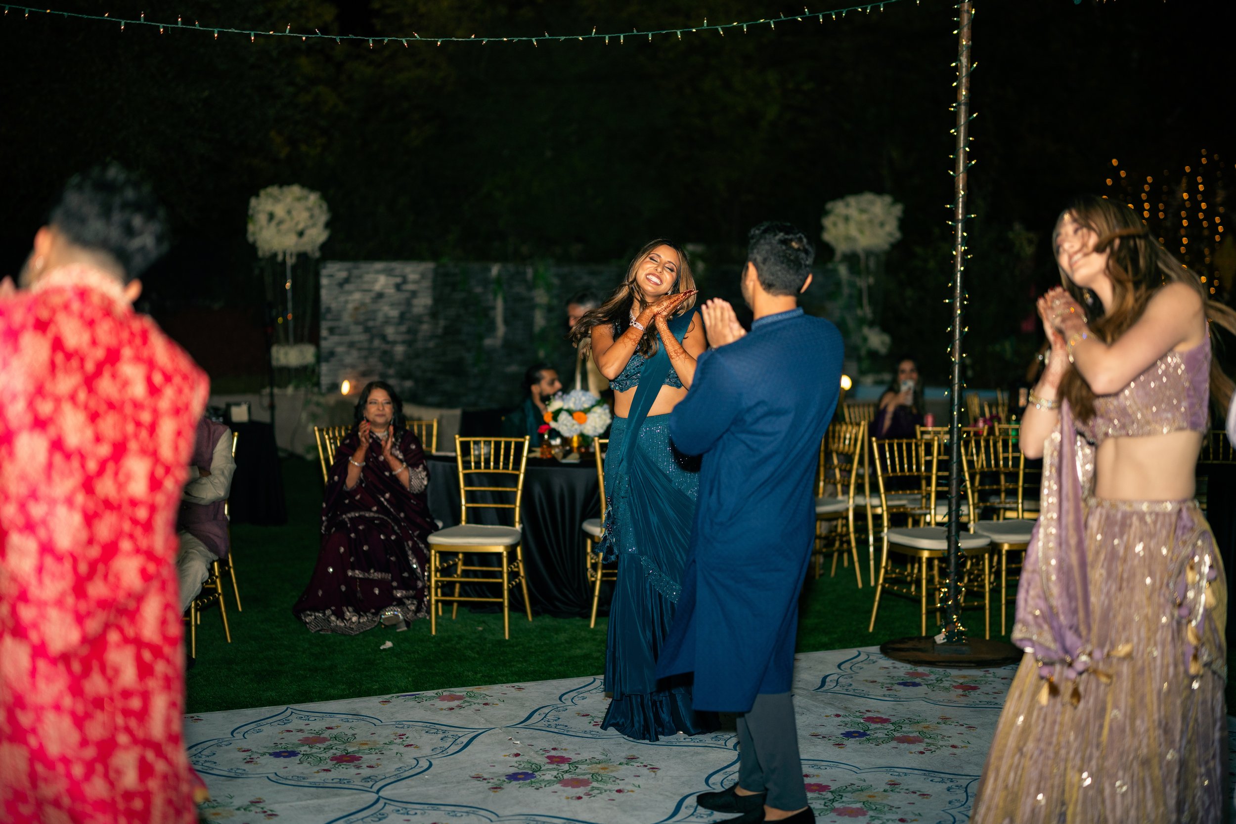 People dancing and celebrating at an outdoor event, possibly a wedding reception, at night. A woman in a blue outfit is smiling and dancing with a man in a blue shirt. Other guests are seated at decorated tables with gold chairs in the background.