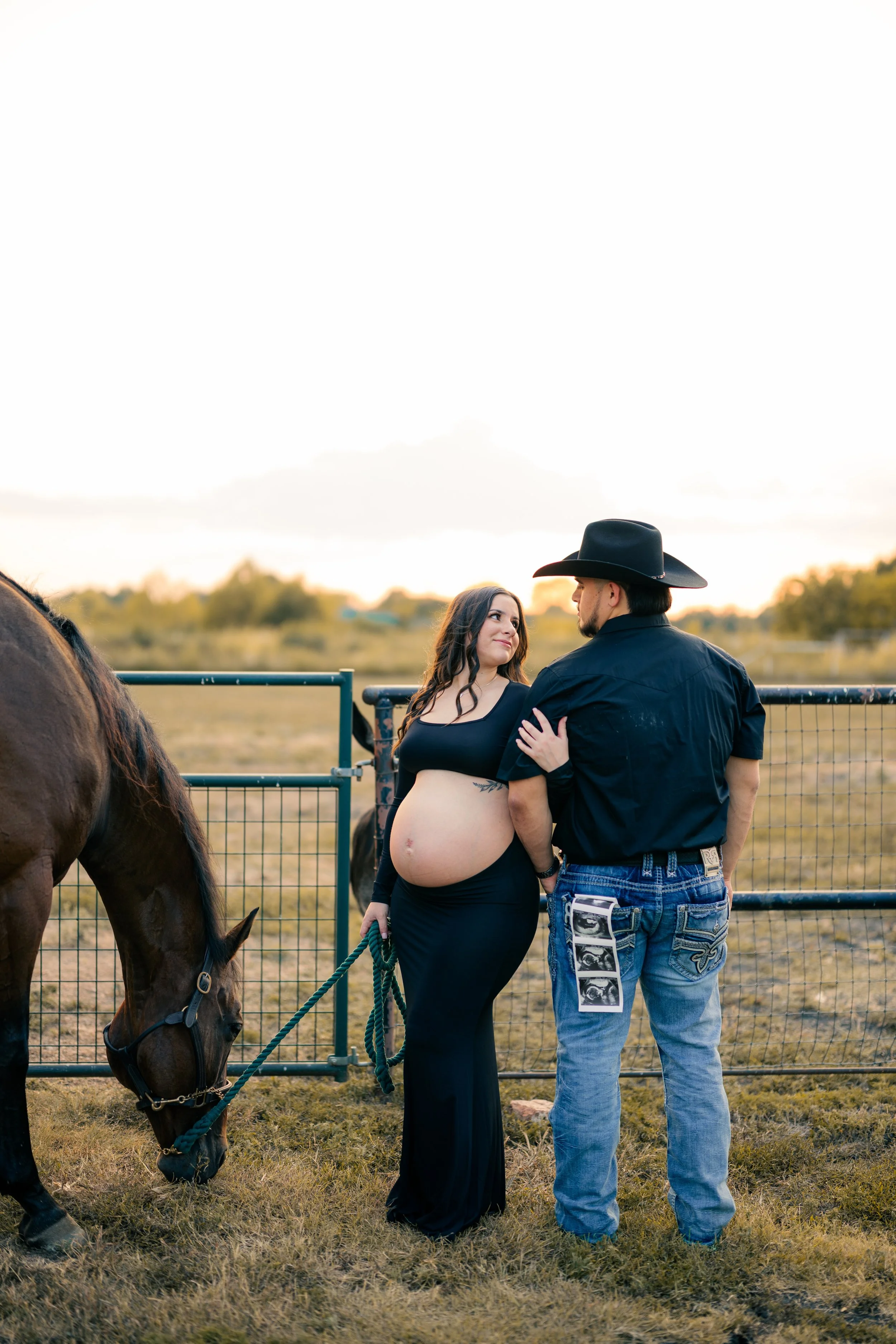A pregnant woman in a black dress holding a rope leash, standing near a horse, looking at a man in a cowboy hat and black shirt, in an outdoor rural setting at sunset.