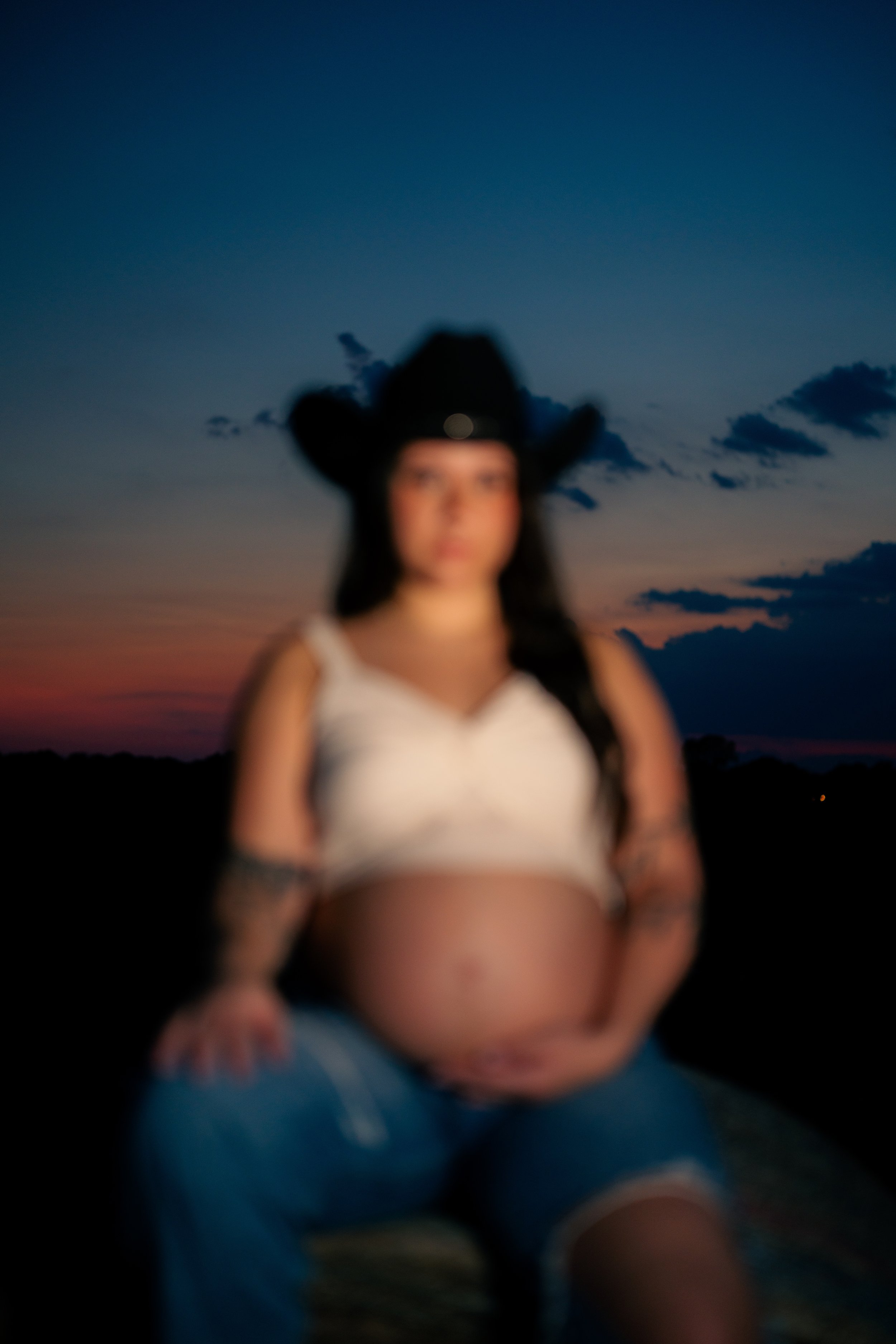 Blurred image of a pregnant woman wearing a black cowboy hat, a white sleeveless top, and blue jeans, sitting outdoors during sunset or dusk.