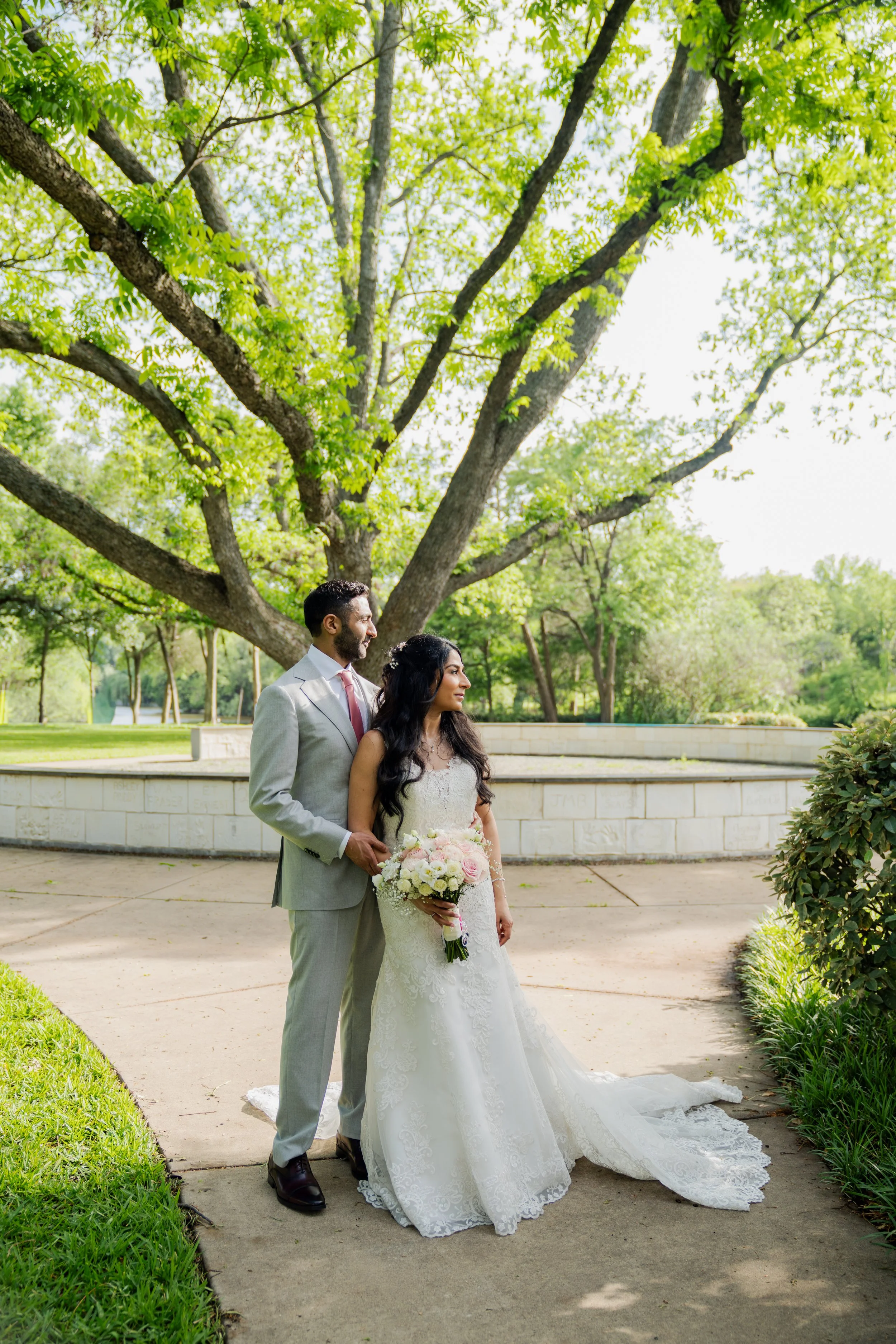 A bridal couple standing on a park pathway under a large leafy tree, with the bride holding a bouquet of white and pink flowers, and the groom in a light gray suit. The bride is in a white wedding dress with lace details, and the groom is in a gray s