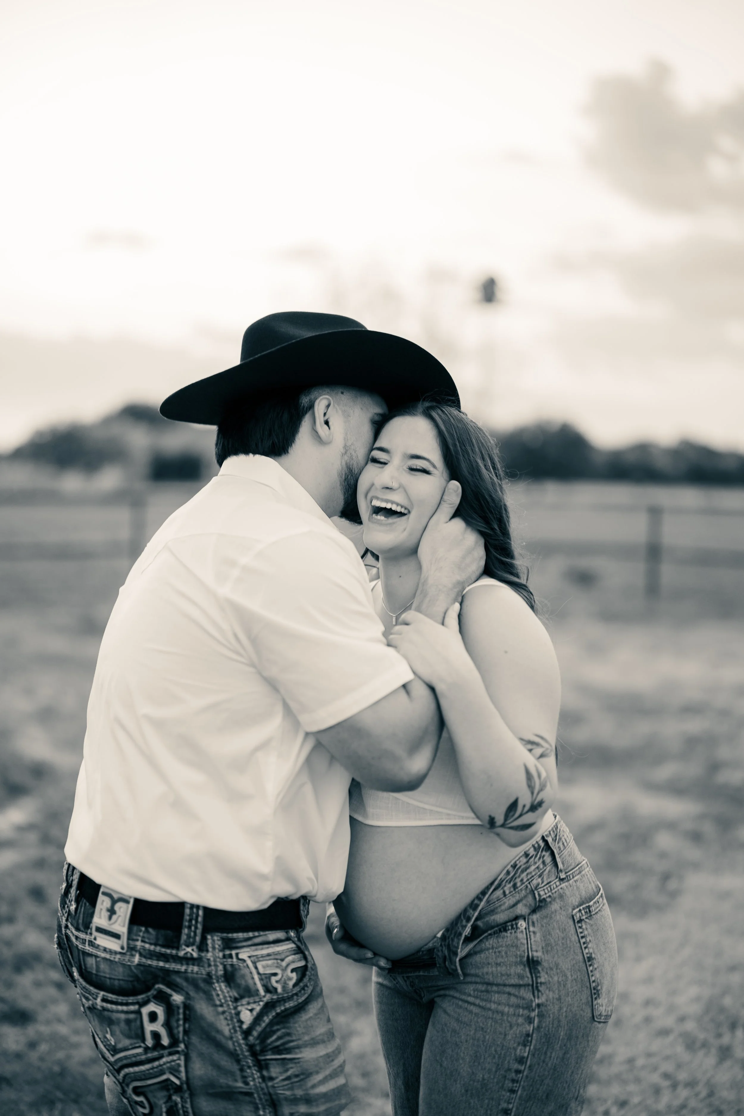 A joyful couple, with the man wearing a cowboy hat and the woman showing a baby bump, sharing a happy moment outdoors.