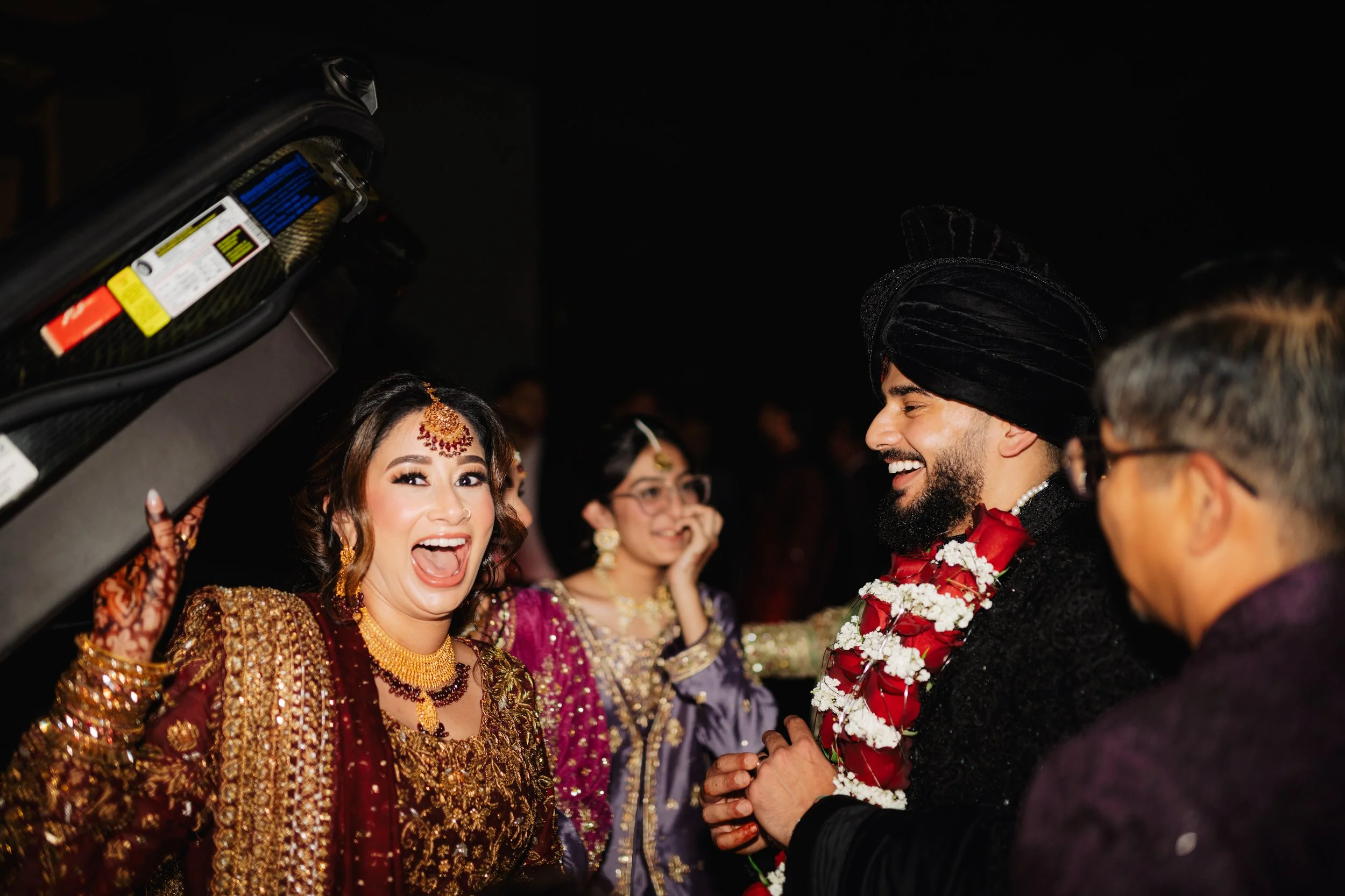 A woman in traditional South Asian attire and jewelry smiles joyfully during a wedding ceremony, holding her hand up in excitement, with men in traditional wedding attire and a woman in glasses in the background.