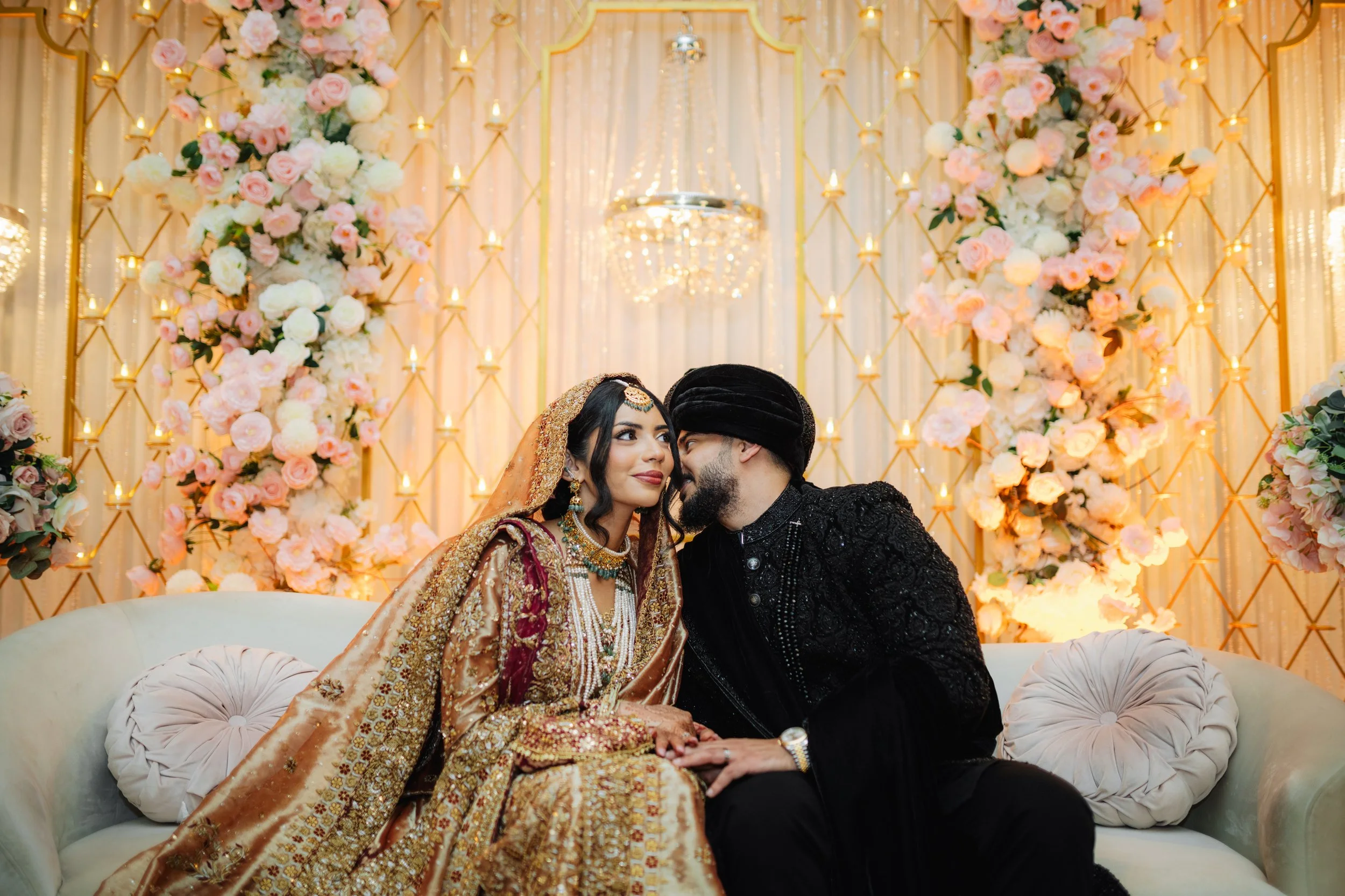 A bride and groom in traditional South Asian wedding attire sitting on a cream-colored sofa with floral arrangements and candles in the background.