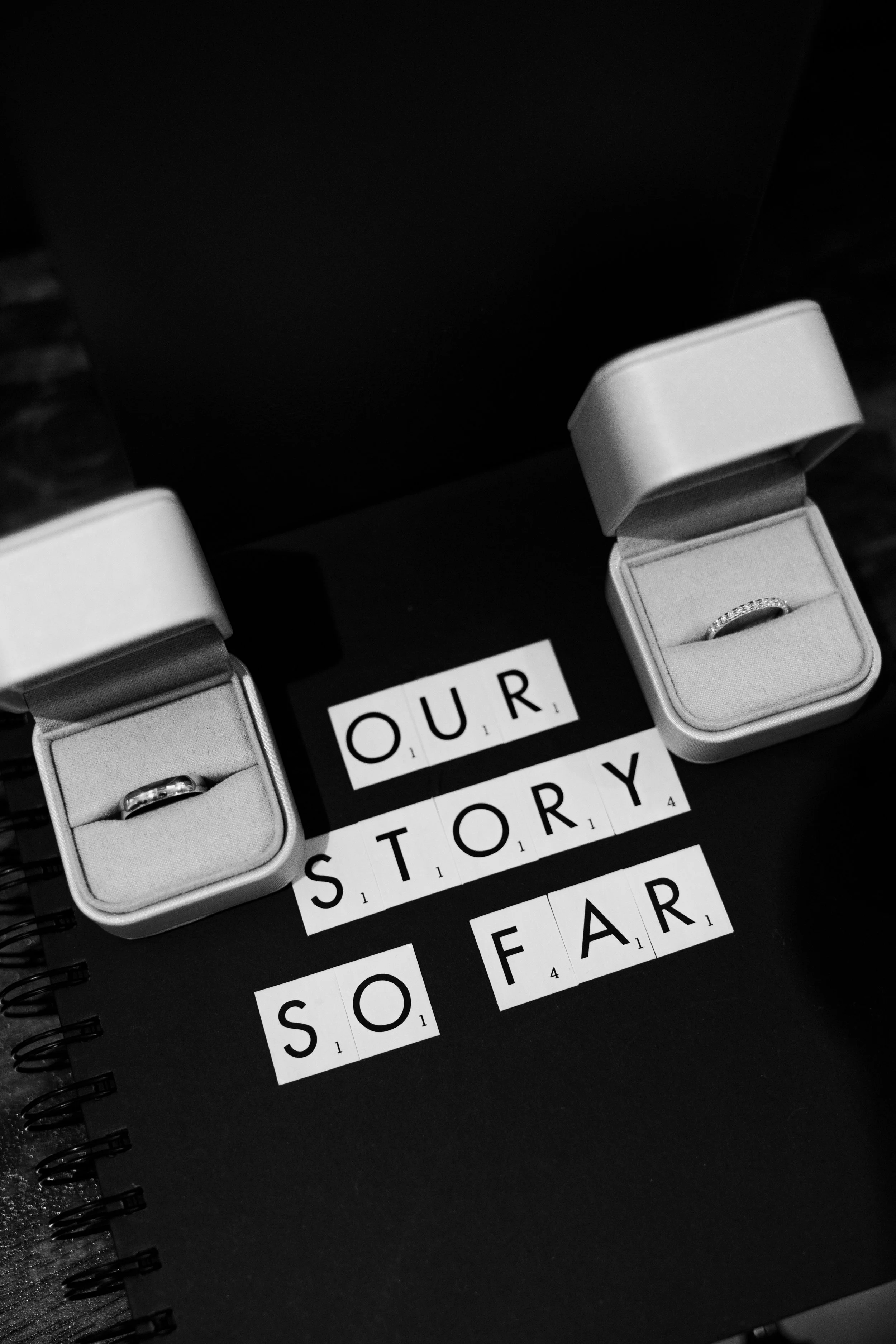 Open jewelry boxes with rings in black notebooks, words spelled out on letter tiles reading 'OUR STORY SO FAR' on a black surface.
