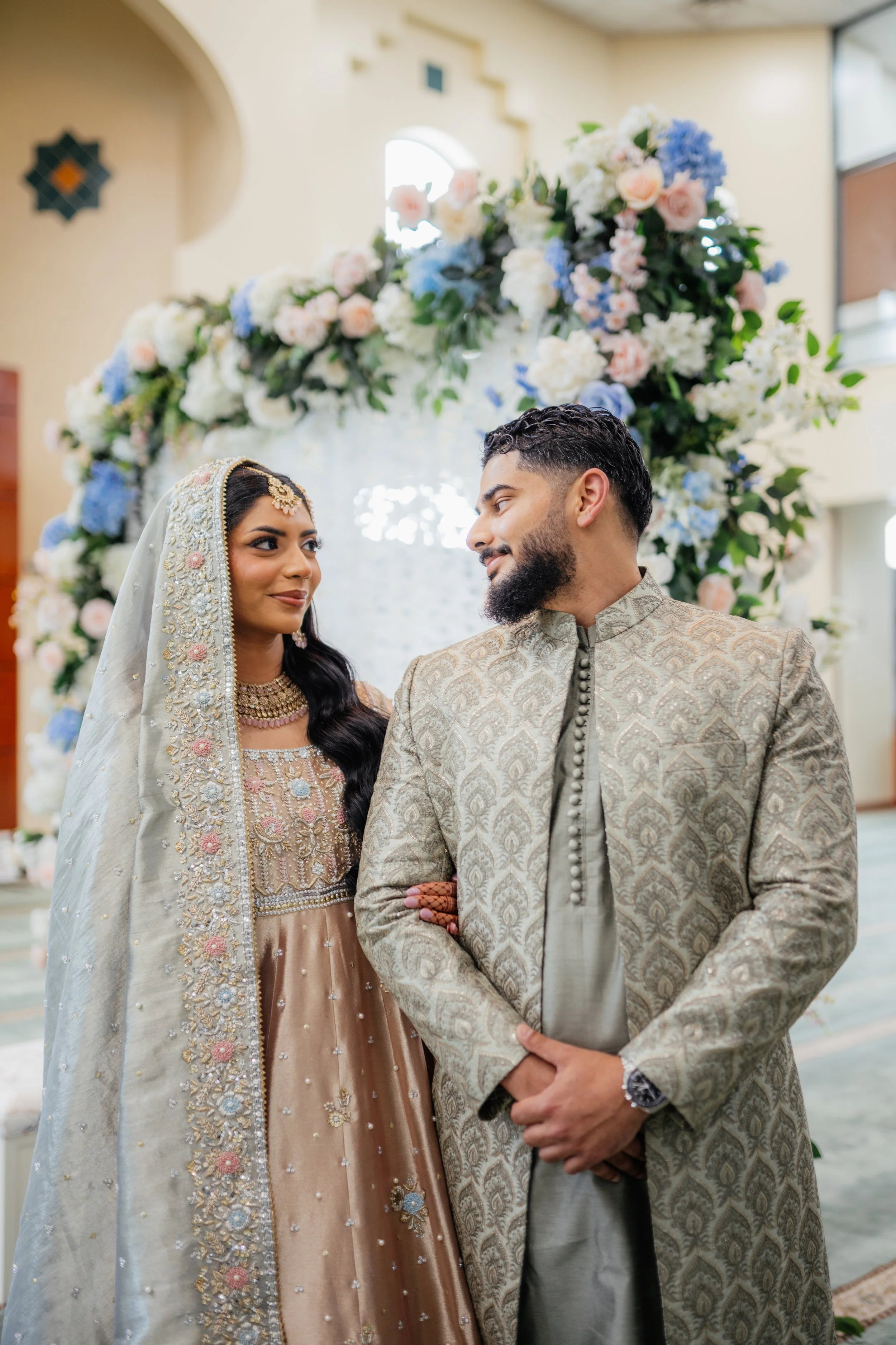 A newlywed couple in traditional Indian wedding attire stands before a floral backdrop, gazing at each other in a brightly lit room.