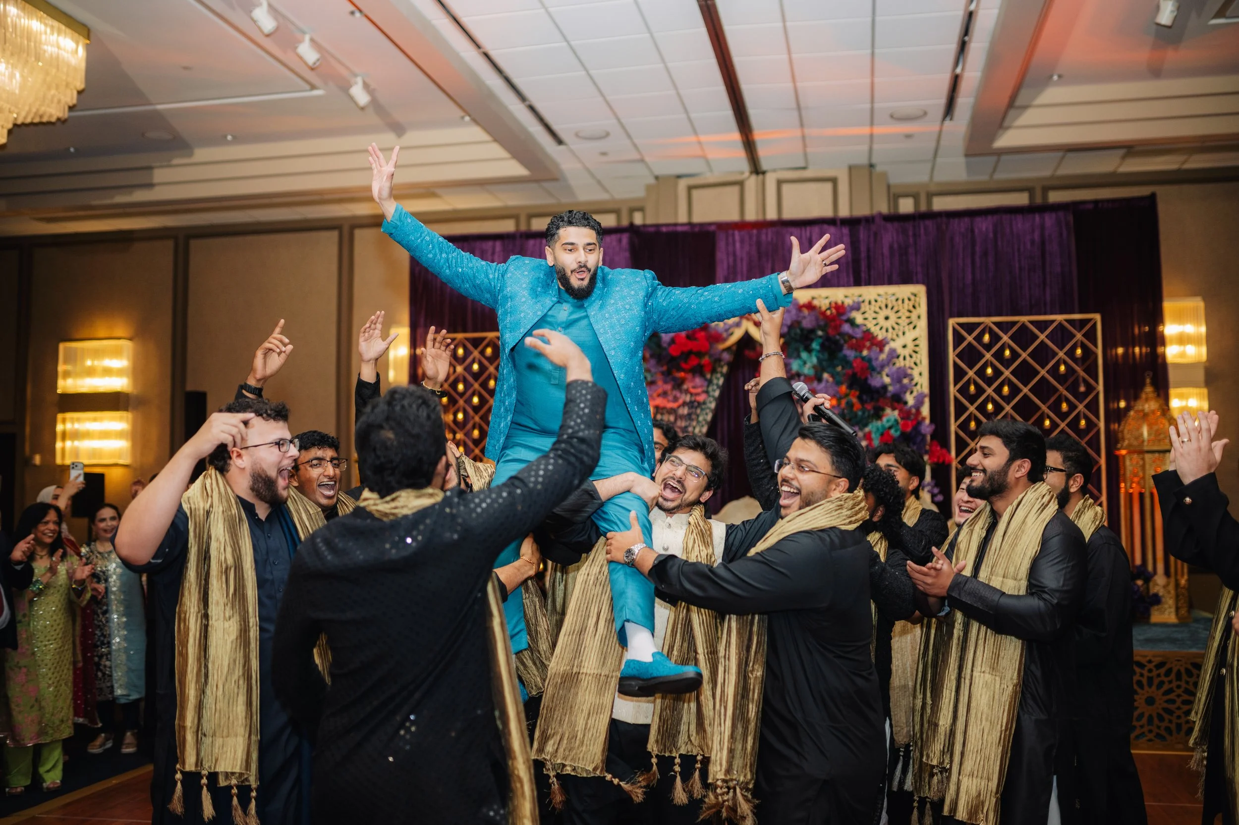 A man in a blue traditional outfit is being lifted by a group of people at a celebration or wedding, surrounded by dancing and joyful expressions in an indoor venue with decorated backdrop.