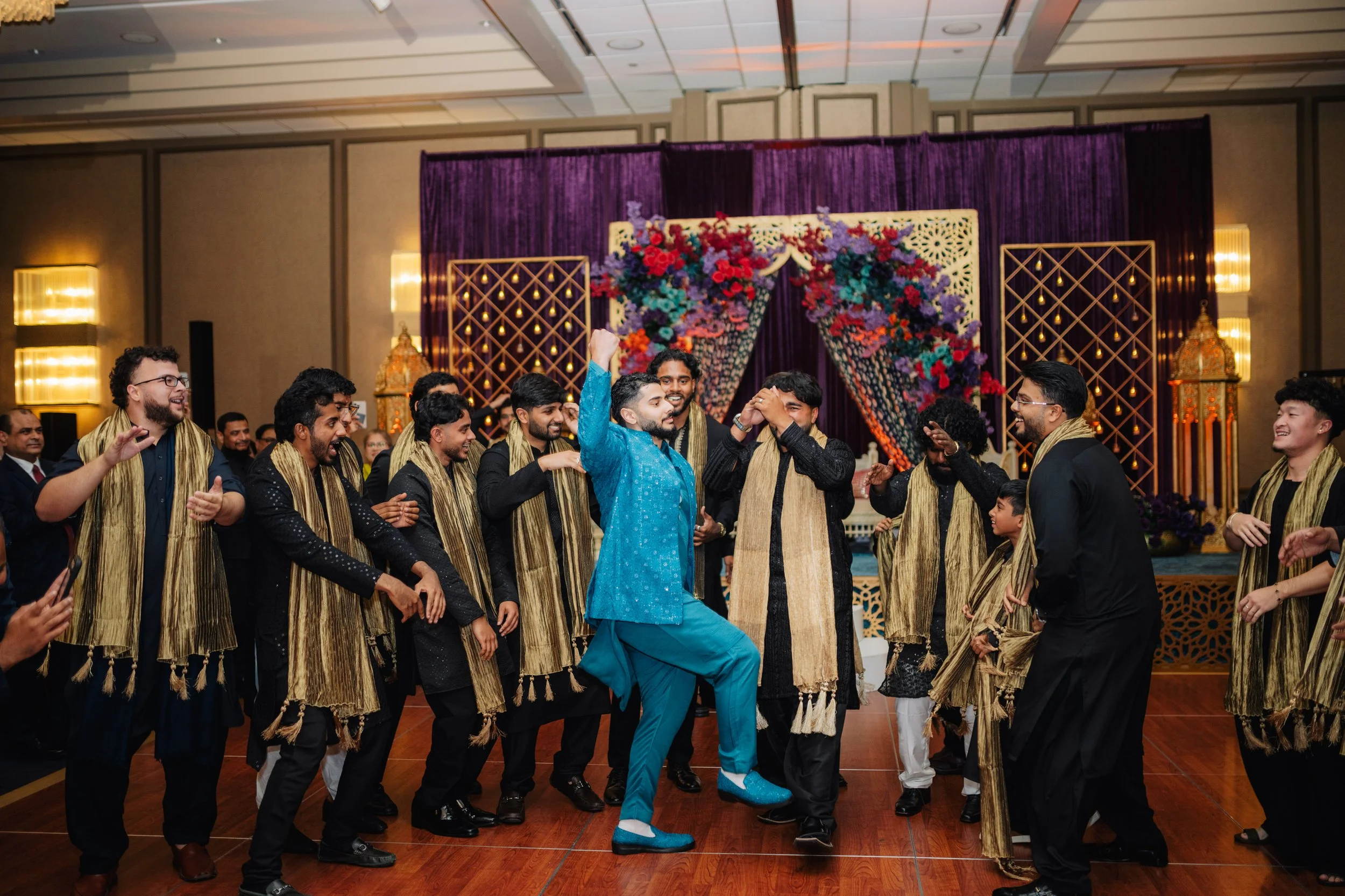A group of men at a wedding celebration dancing in traditional Indian attire, with a decorated stage in the background featuring purple drapes and floral arrangements.