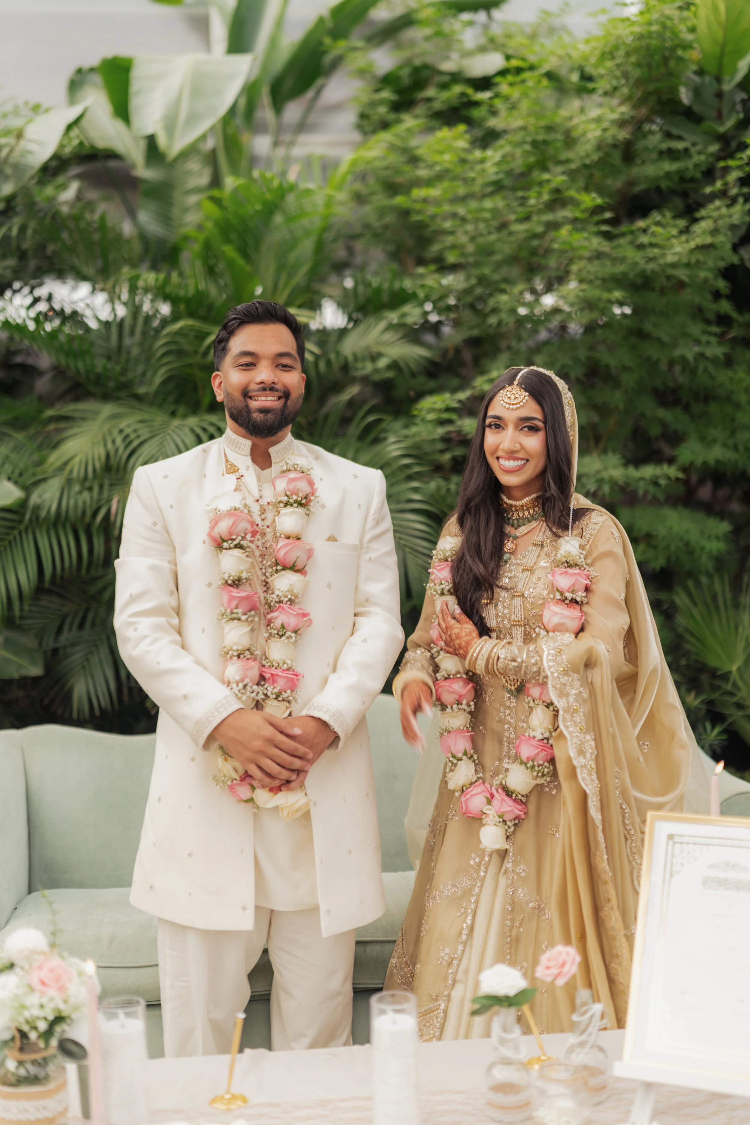 A newly married Indian couple dressed in traditional wedding attire standing indoors with greenery behind them. They are smiling, wearing flower garlands, and looking happy.