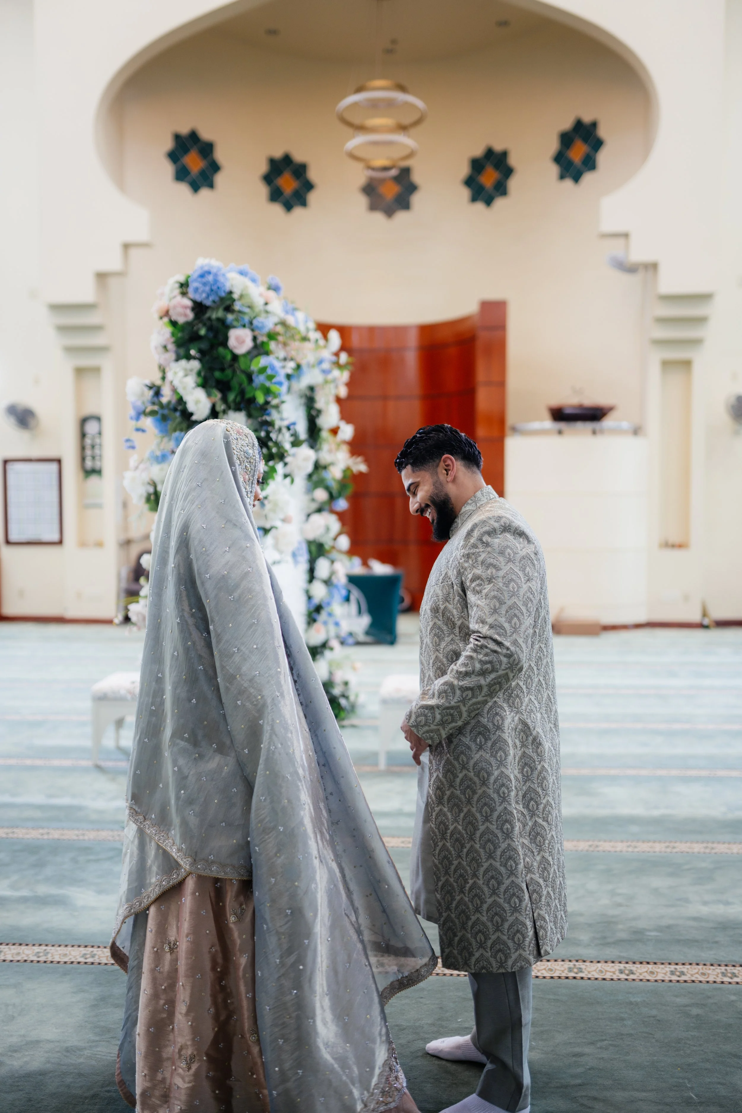 A bride and groom in traditional South Asian wedding attire standing indoors facing each other with a floral arch behind them.