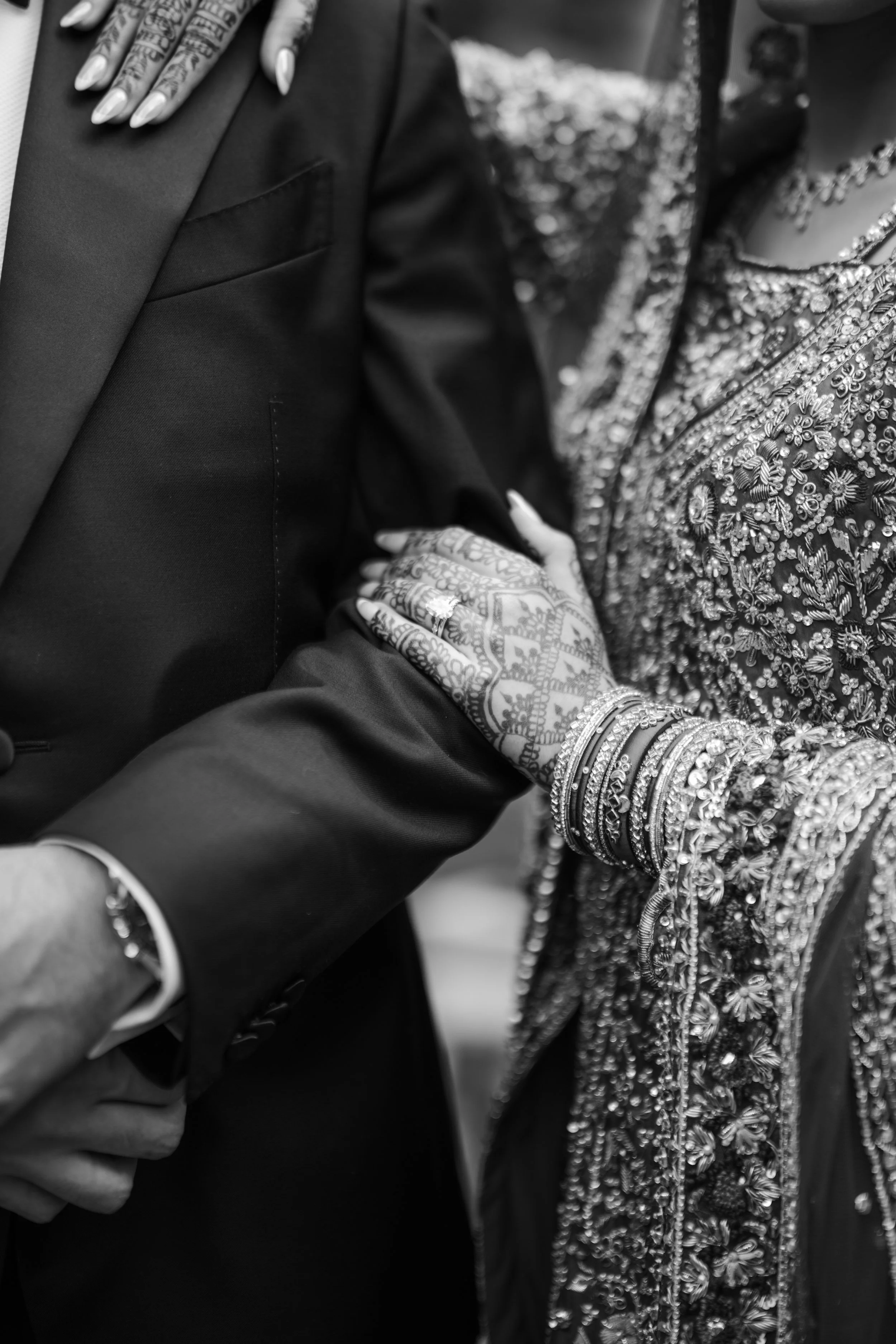 A close-up black and white photograph of Indian wedding attire, showing a man in a dark suit with ornate mehndi designs on his hand, and a woman in an elaborately embroidered traditional outfit with numerous bangles and jewelry.