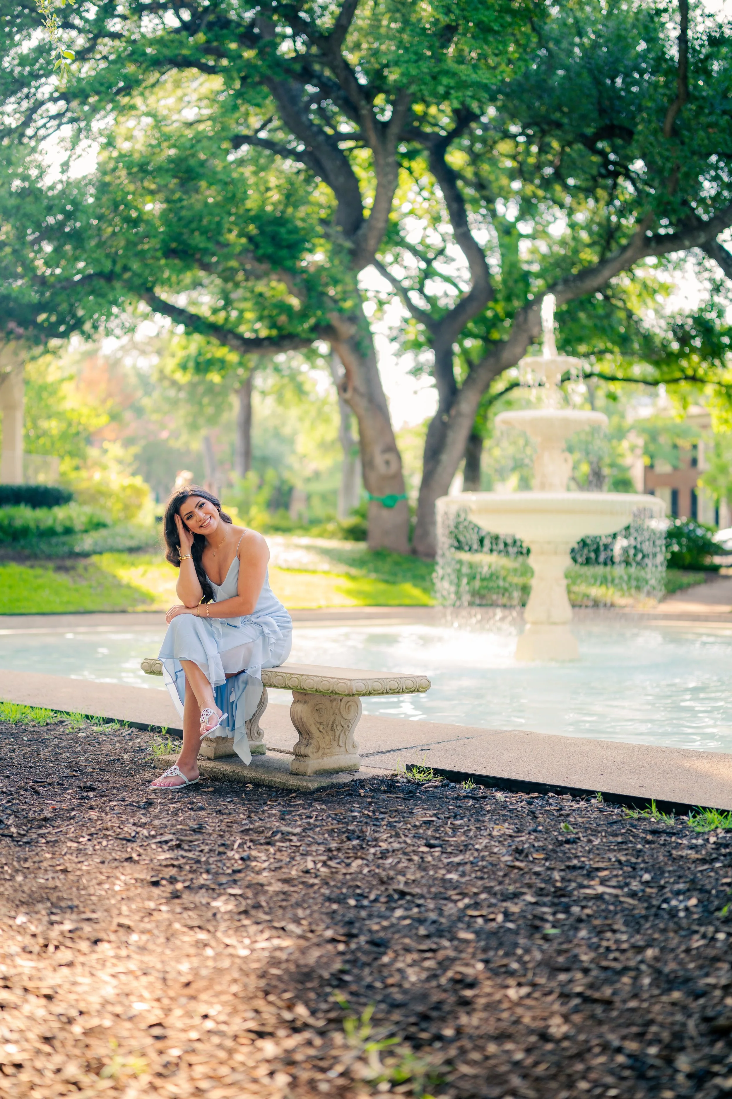 A woman in a light blue dress sitting on a stone bench next to a fountain in a park with large green trees.