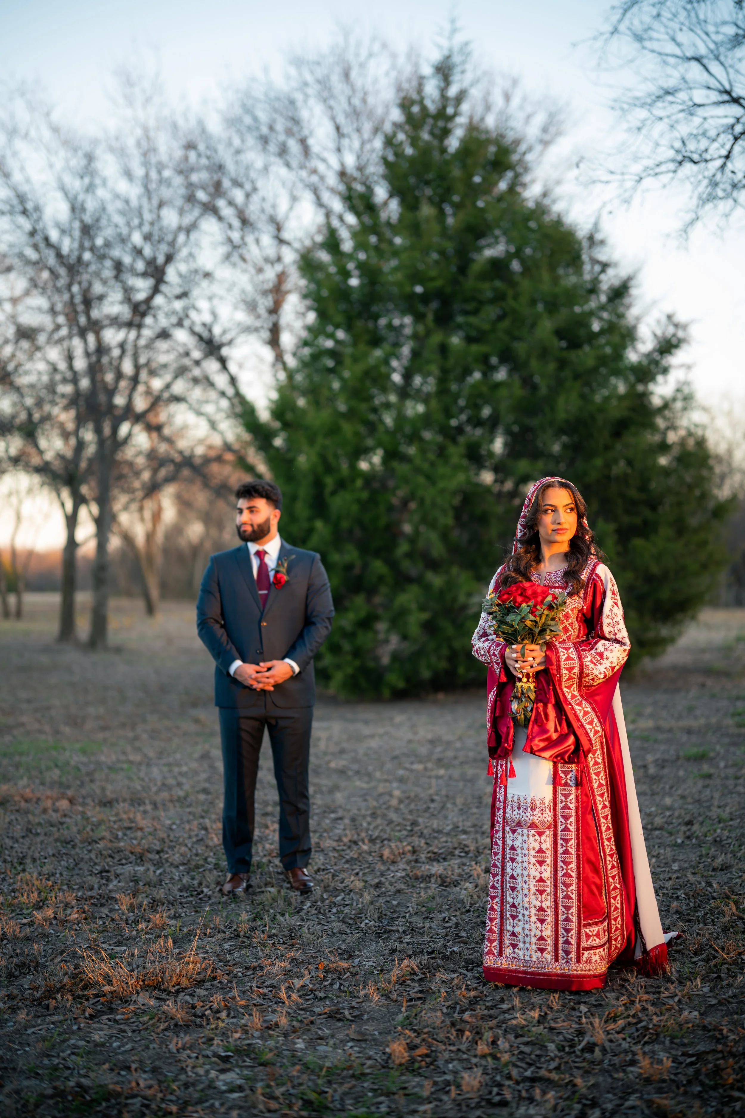 A couple standing outdoors during sunset, with the woman wearing traditional red and white attire and holding a bouquet of red flowers, and the man in a dark suit with a red tie and boutonniere, in front of a large green tree.