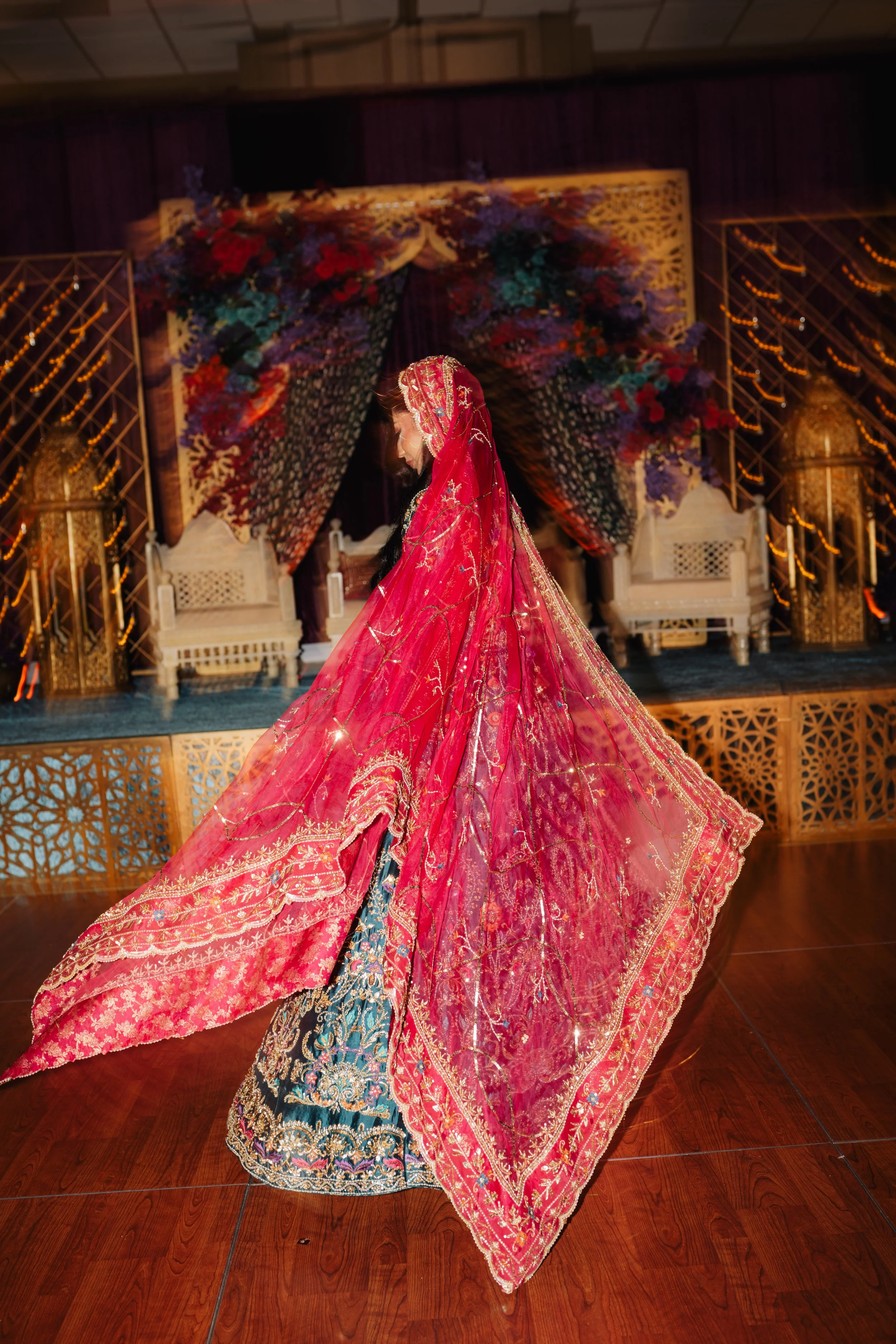 A woman wearing a traditional ornate pink and gold bridal gown and veil, standing on a decorated stage with chairs and floral arrangements in the background.