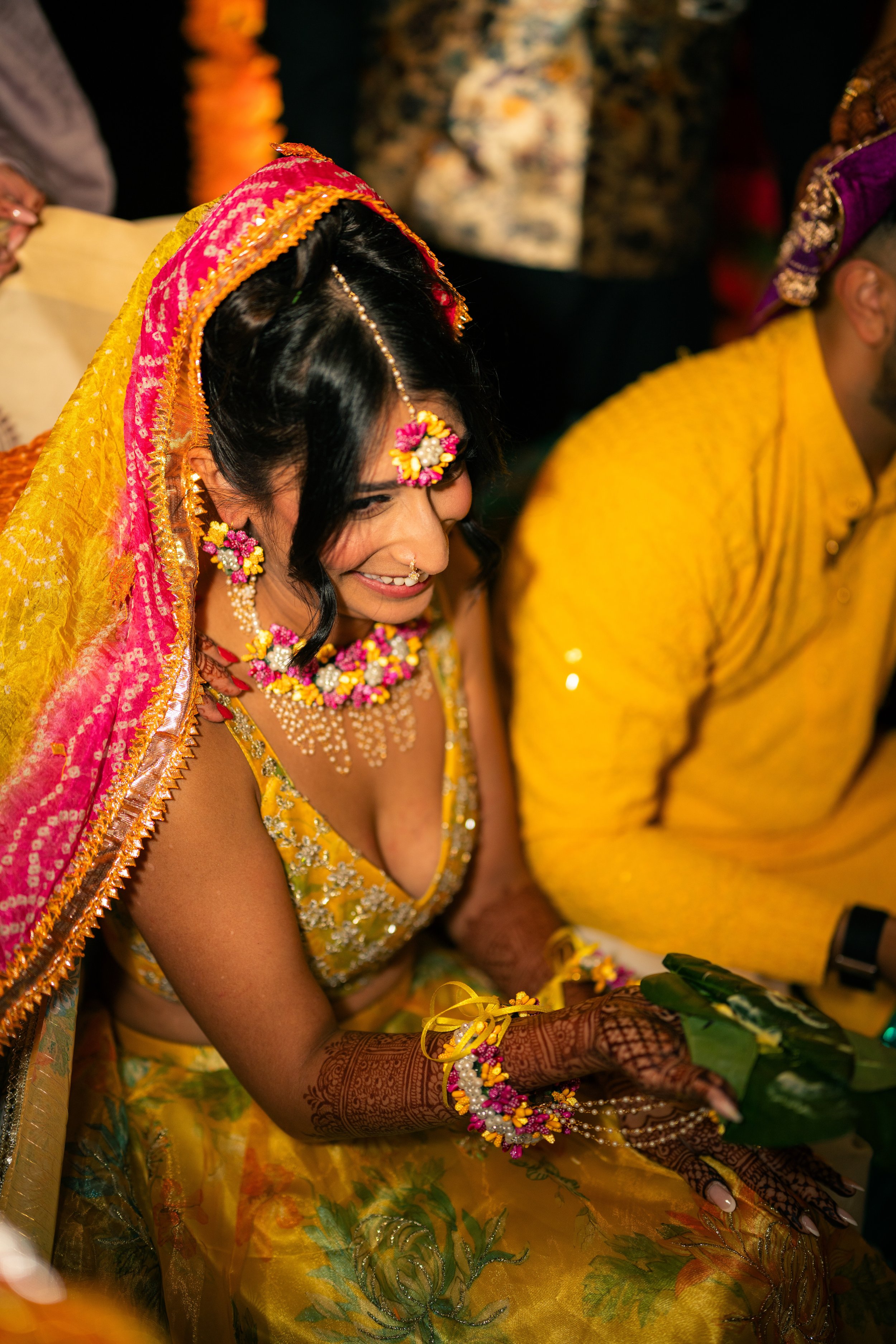A woman dressed in a yellow traditional outfit with floral jewelry, henna on her hands, and a pink and yellow dupatta, smiling during a cultural or wedding celebration.