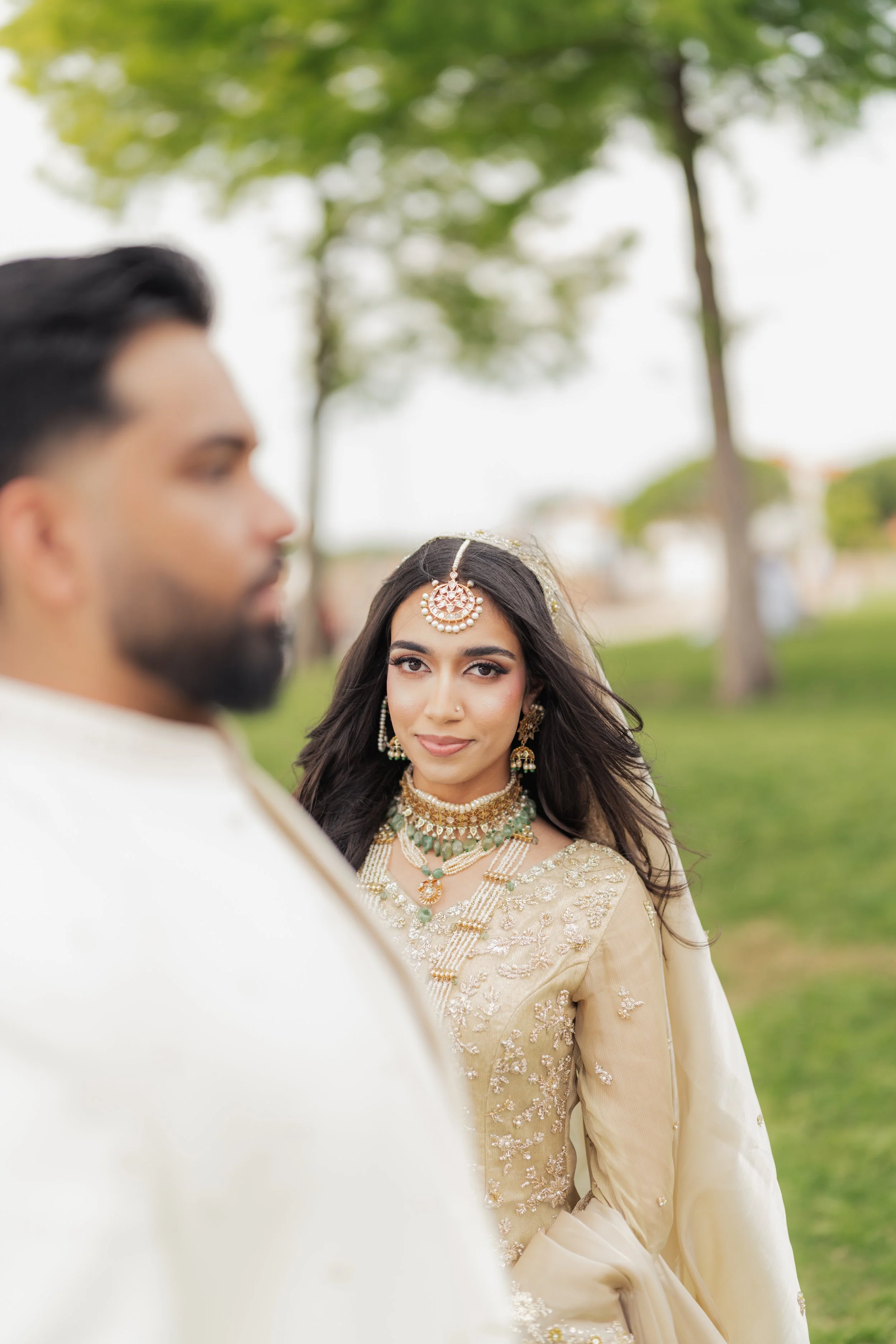 A woman dressed in traditional Indian attire and jewelry looks at the camera, while a man in a white outfit stands in the foreground blurred, outdoors with trees and sky in the background.