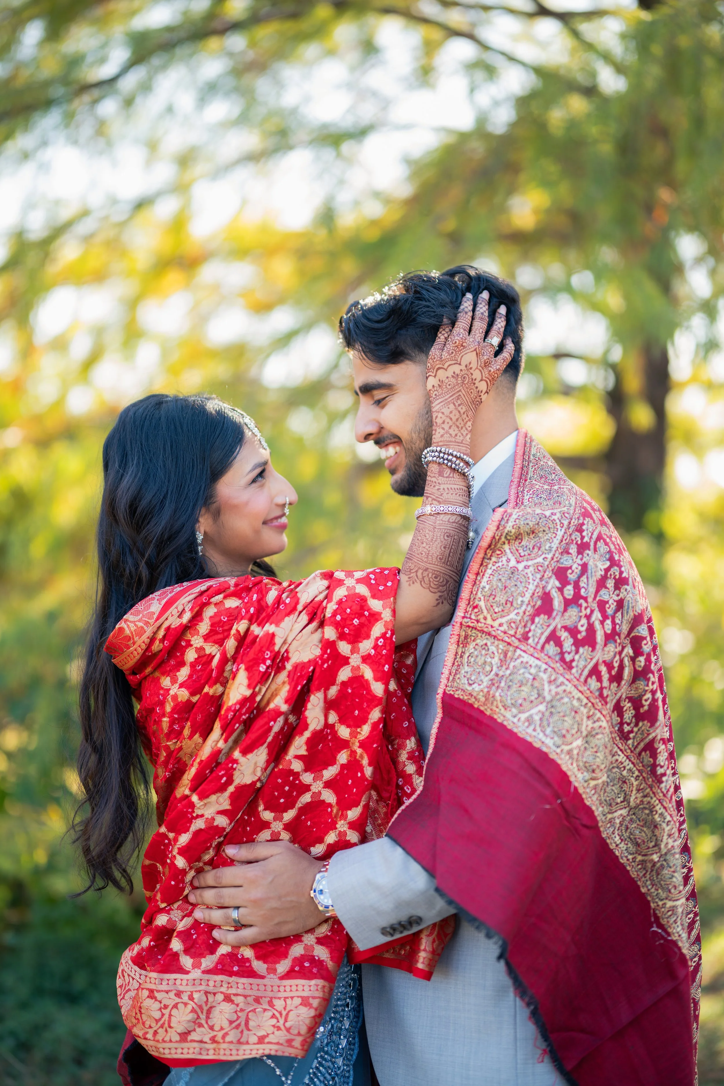 A couple dressed in traditional South Asian attire, gazing at each other lovingly outdoors with autumn trees in the background.