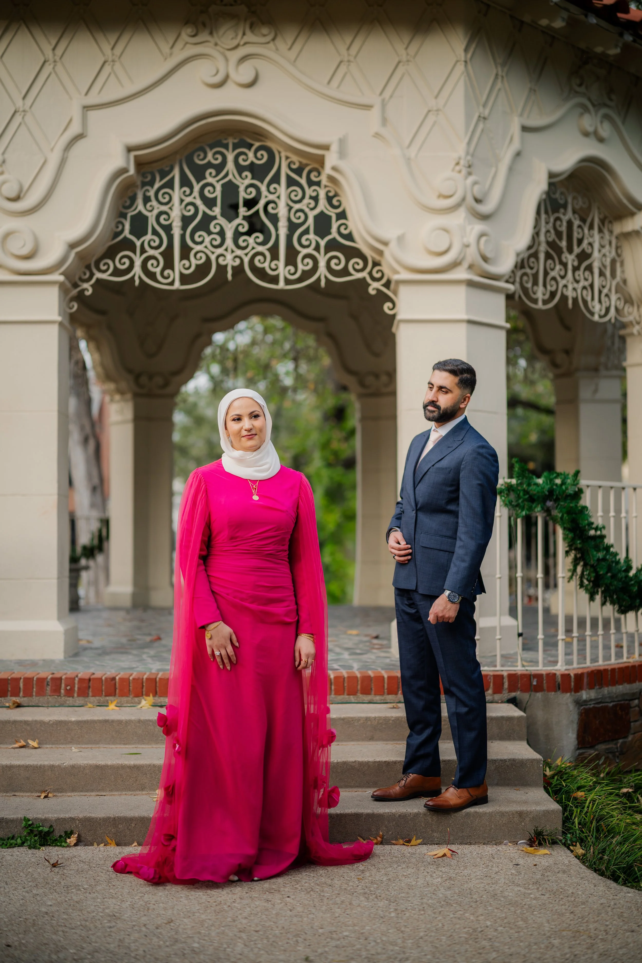 A woman in a bright pink dress and a white hijab standing next to a man in a navy suit outside a decorative, ornate building archway with greenery in the background.