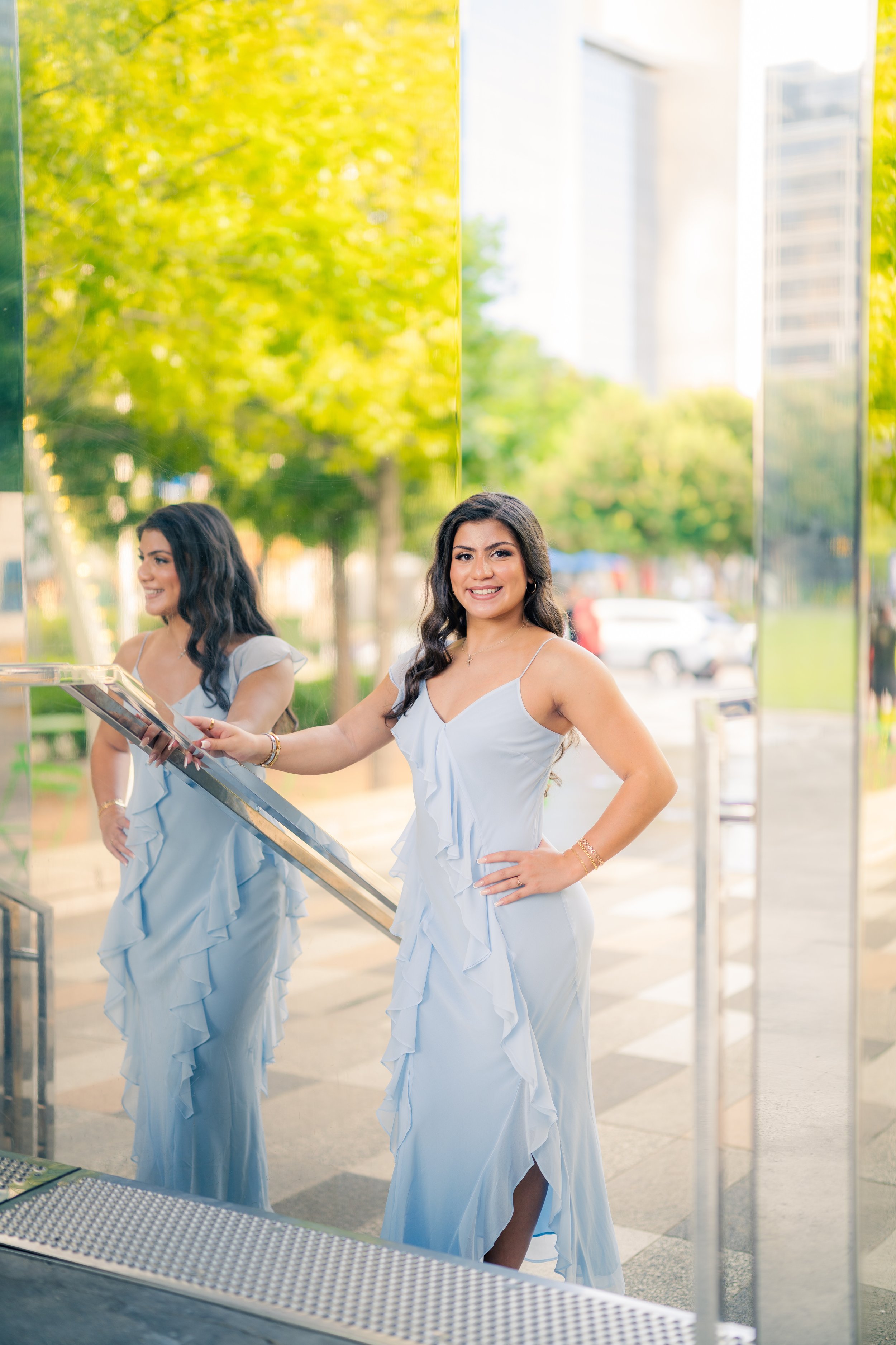 A woman in a light blue ruffled dress standing outside on a sunny day, smiling, with her reflection visible in a large glass surface behind her.