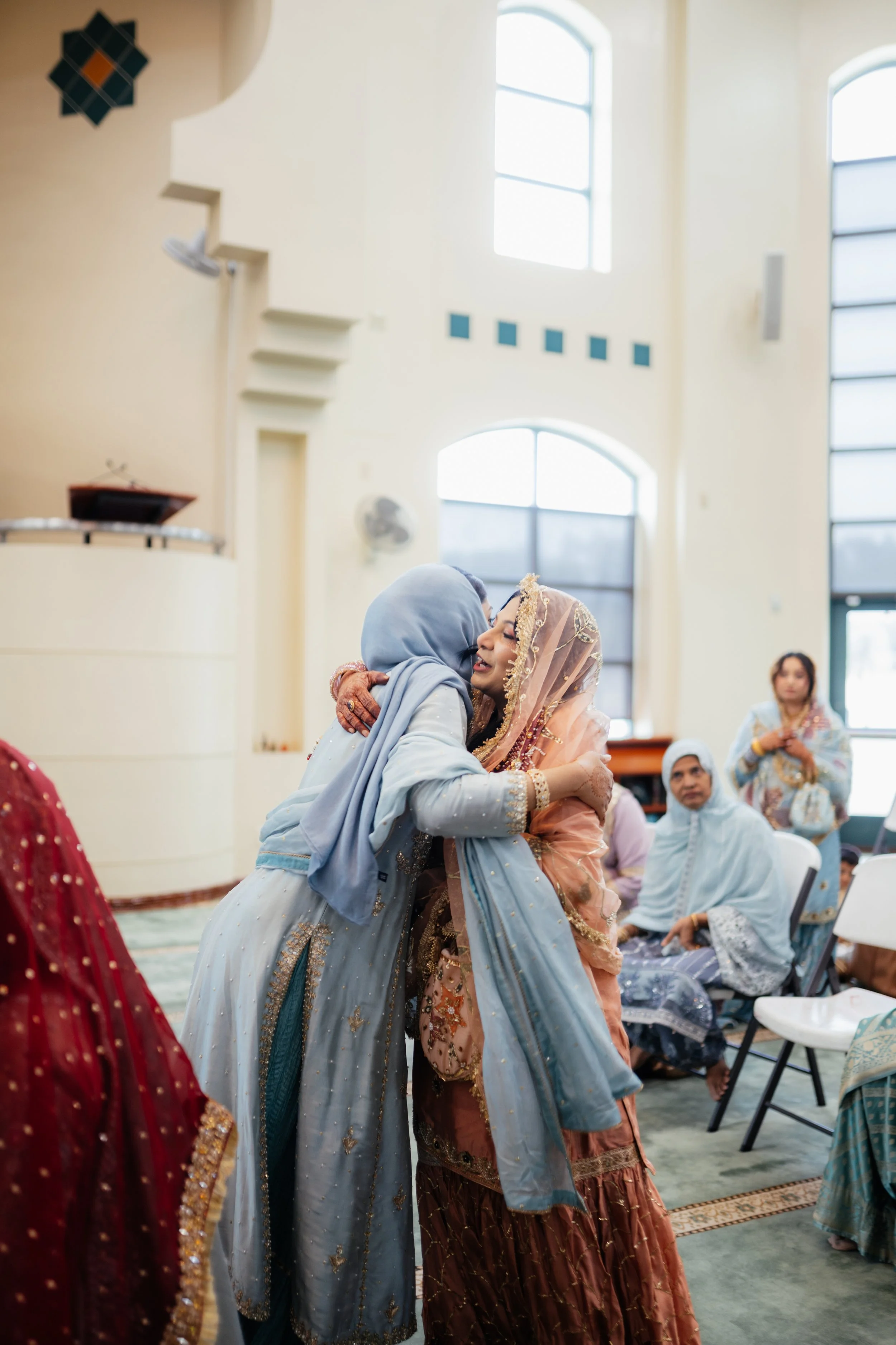 Women in colorful traditional dresses hugging inside a brightly lit room with large windows.