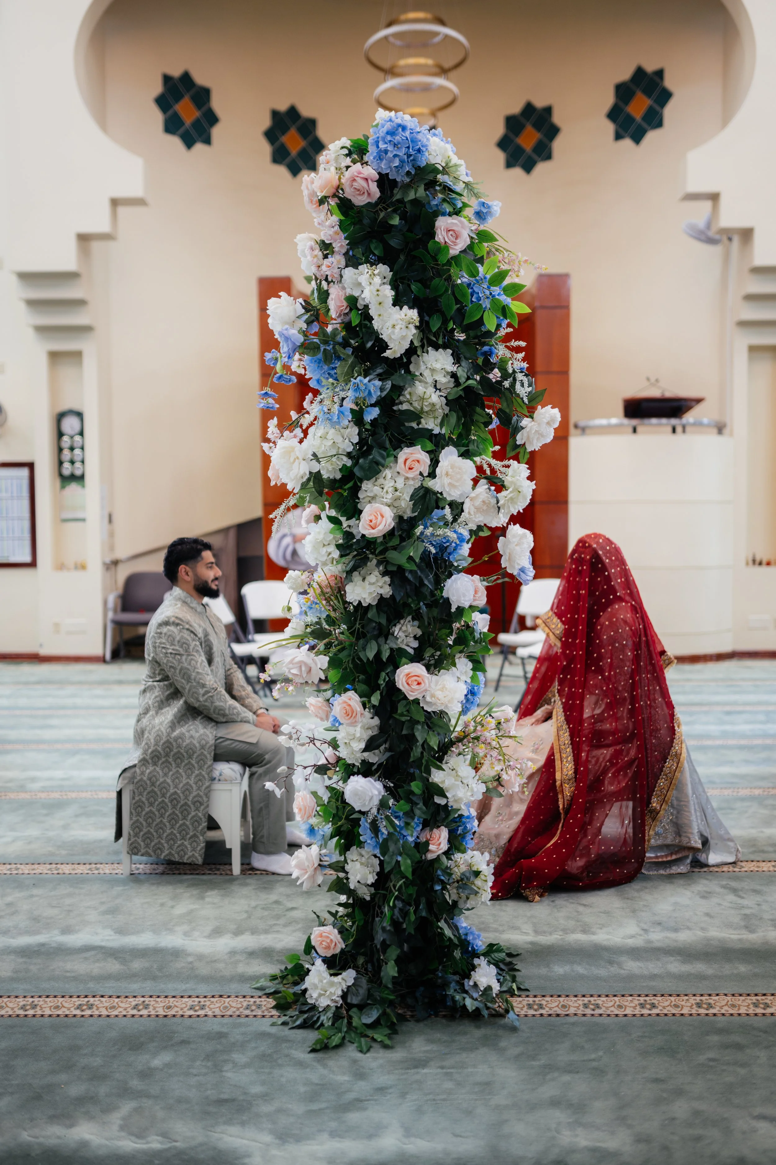 A traditional wedding ceremony taking place indoors, with a floral arch in the center decorated with white, pink, and blue flowers. A man in a silver patterned suit is kneeling on one side, facing a woman in a red and gold sari who is also kneeling. 