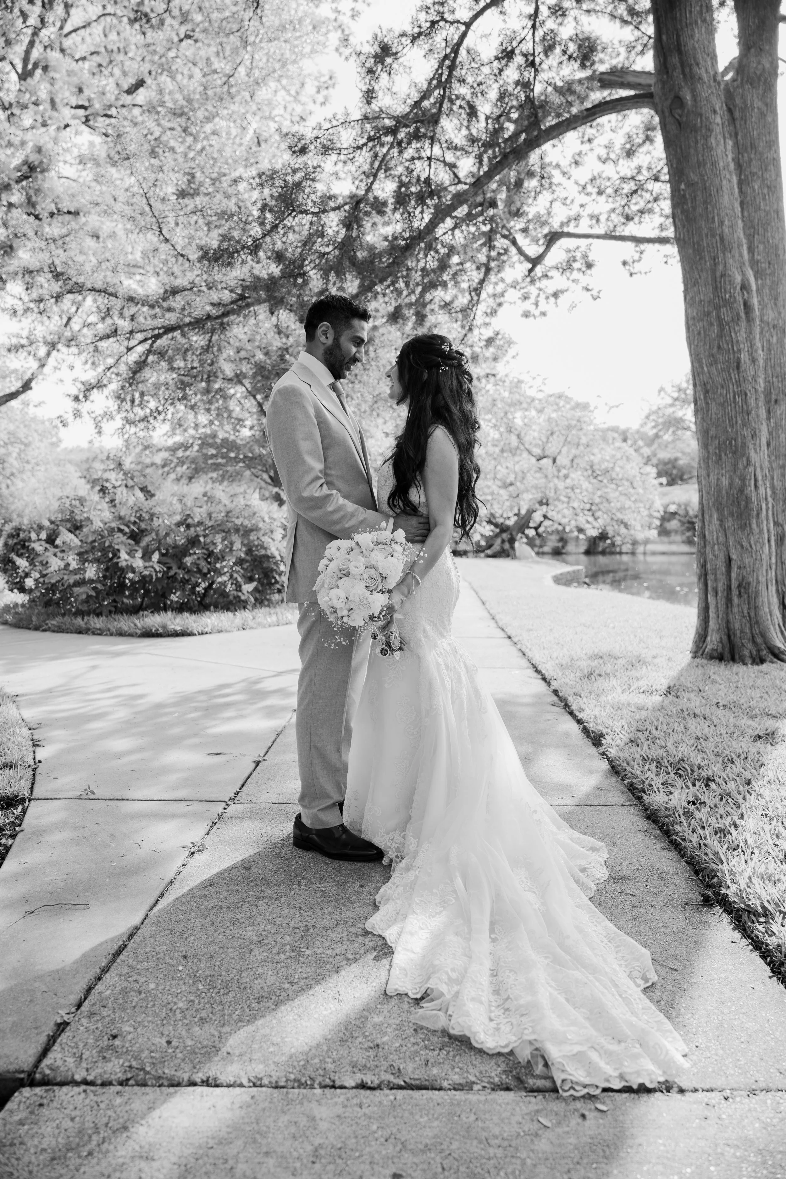 A black and white photo of a bride and groom standing close together on a sidewalk in a park, holding hands and gazing into each other's eyes, surrounded by trees and sunlight.