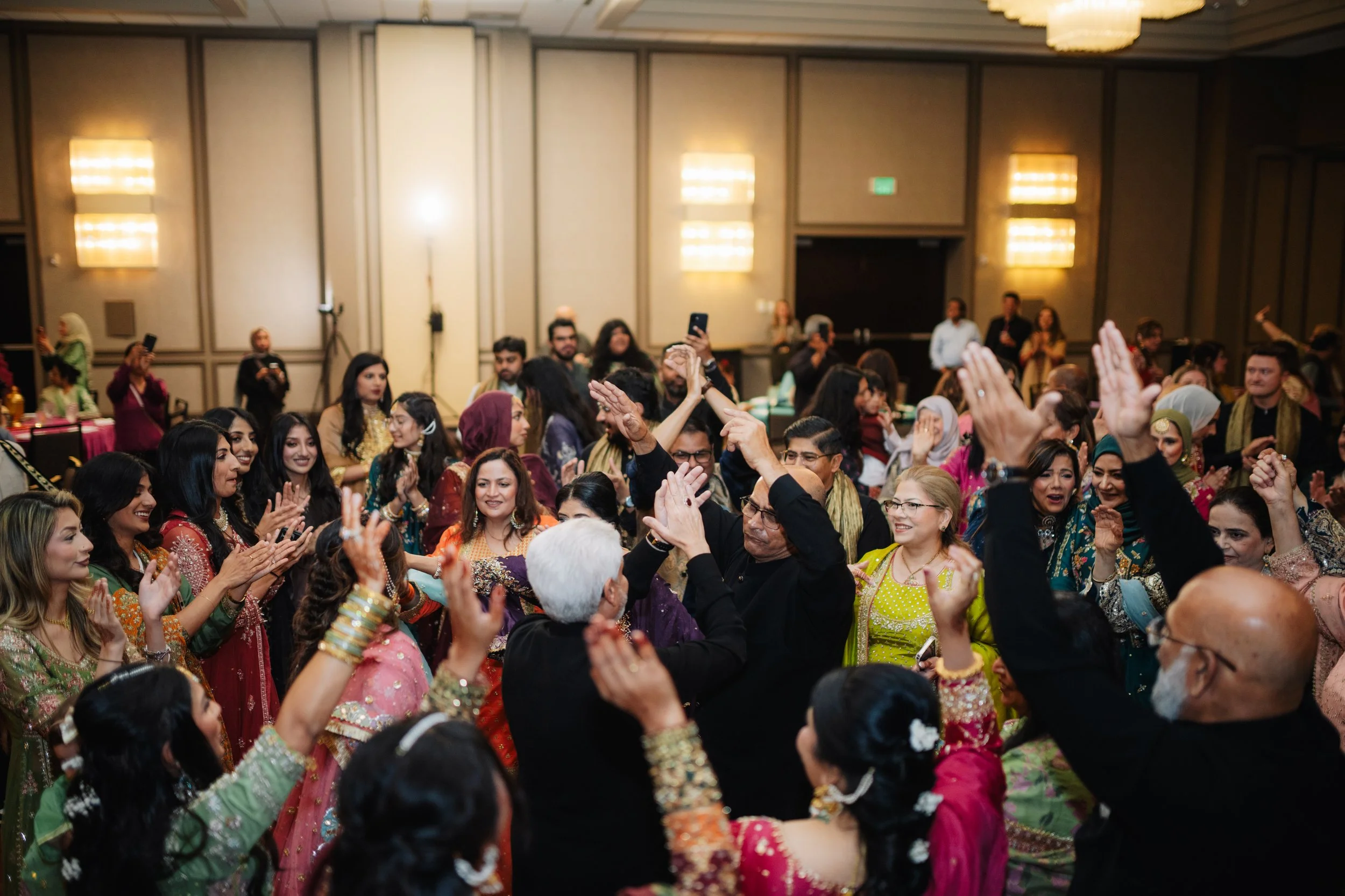 A large group of people at an indoor celebration, wearing colorful traditional attire, dancing and enjoying the event.
