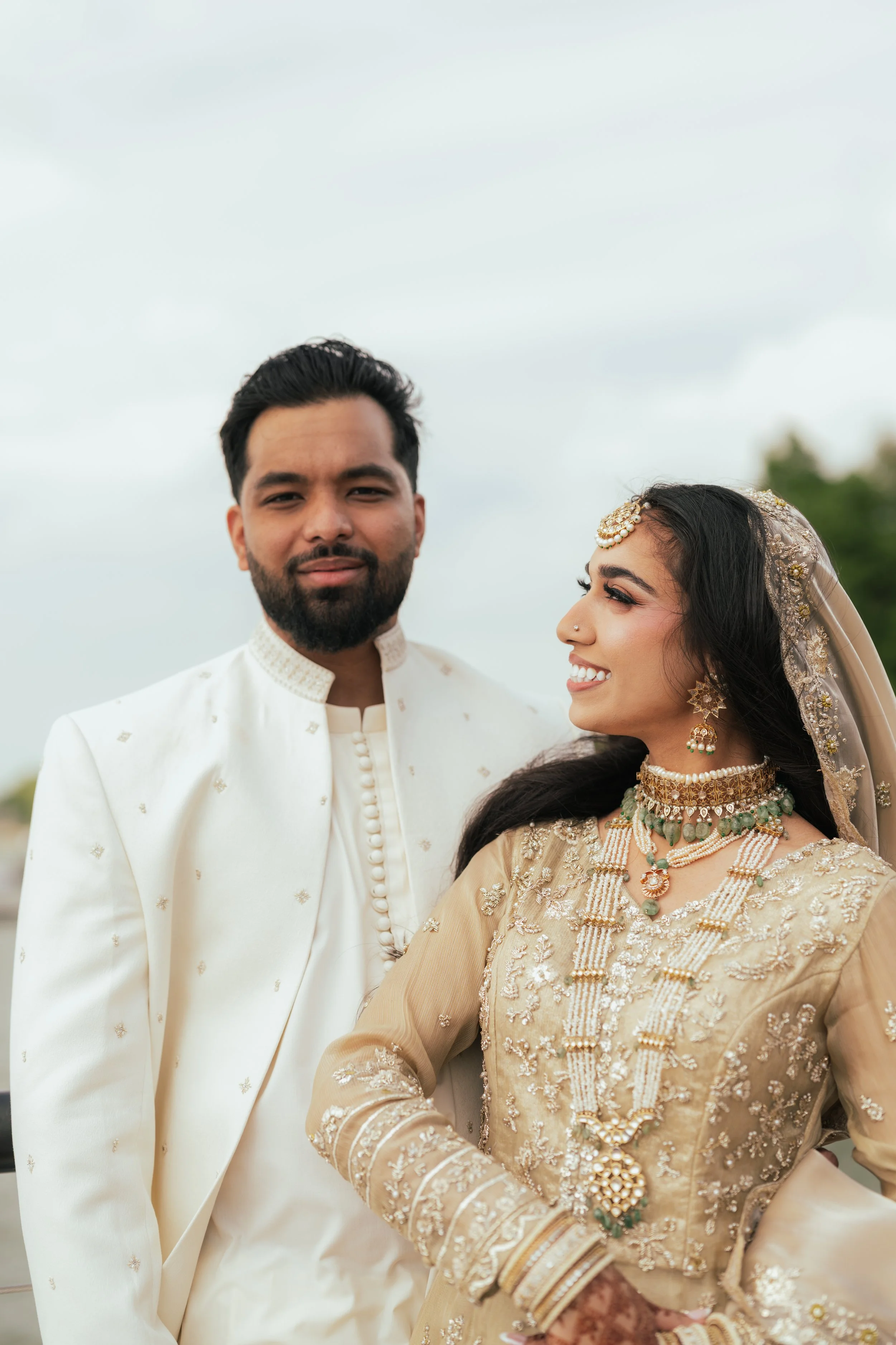 A bride and groom dressed in traditional Indian wedding attire, standing outdoors against a cloudy sky. The bride is smiling and wearing gold jewelry and a beige embroidered outfit, while the groom is wearing a white sherwani with intricate embroider
