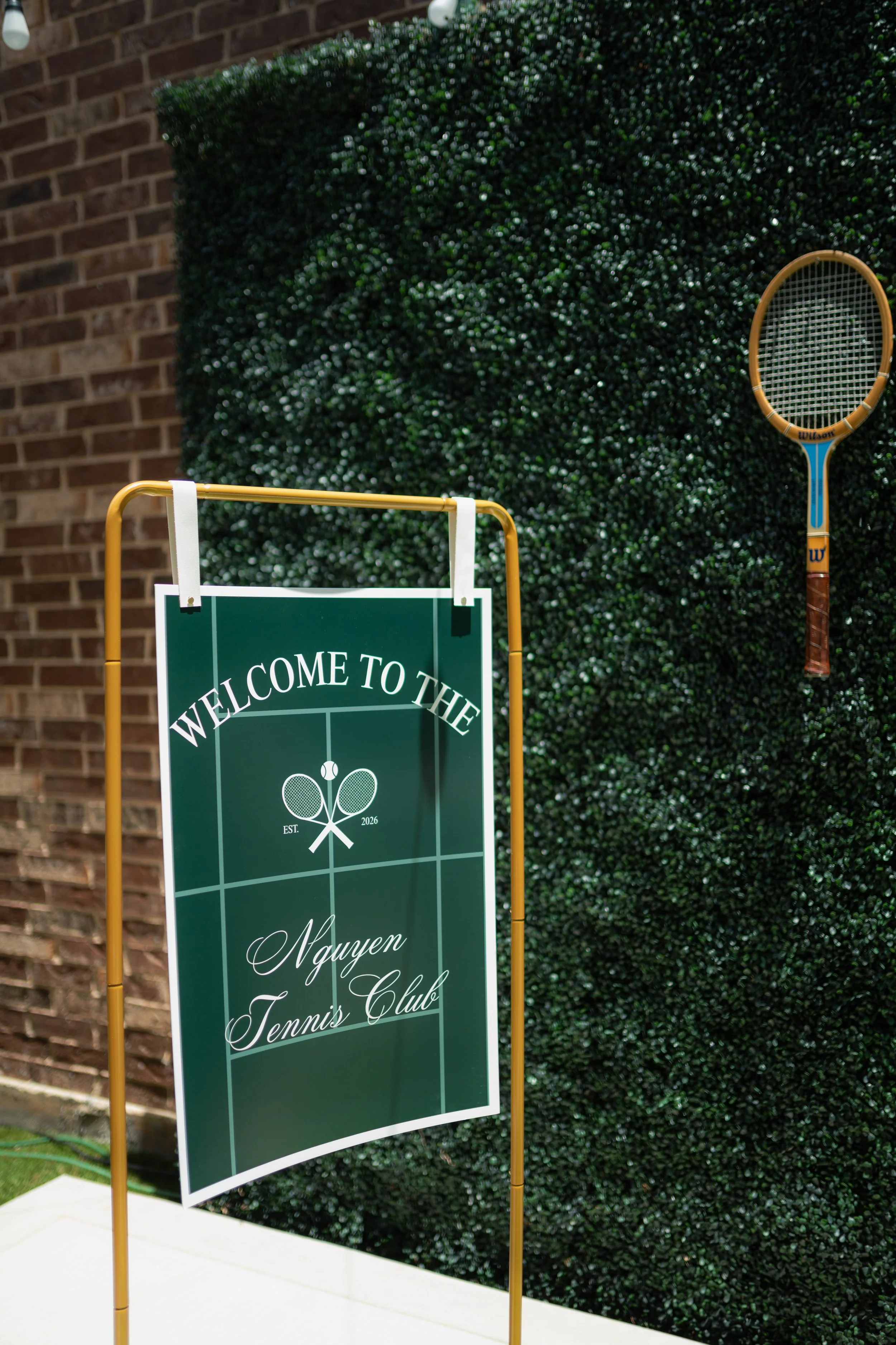 Sign that reads "Welcome to the Nguyen Tennis Club" with a tennis racket hanging on a green hedge background, brick wall, and a yellow frame.