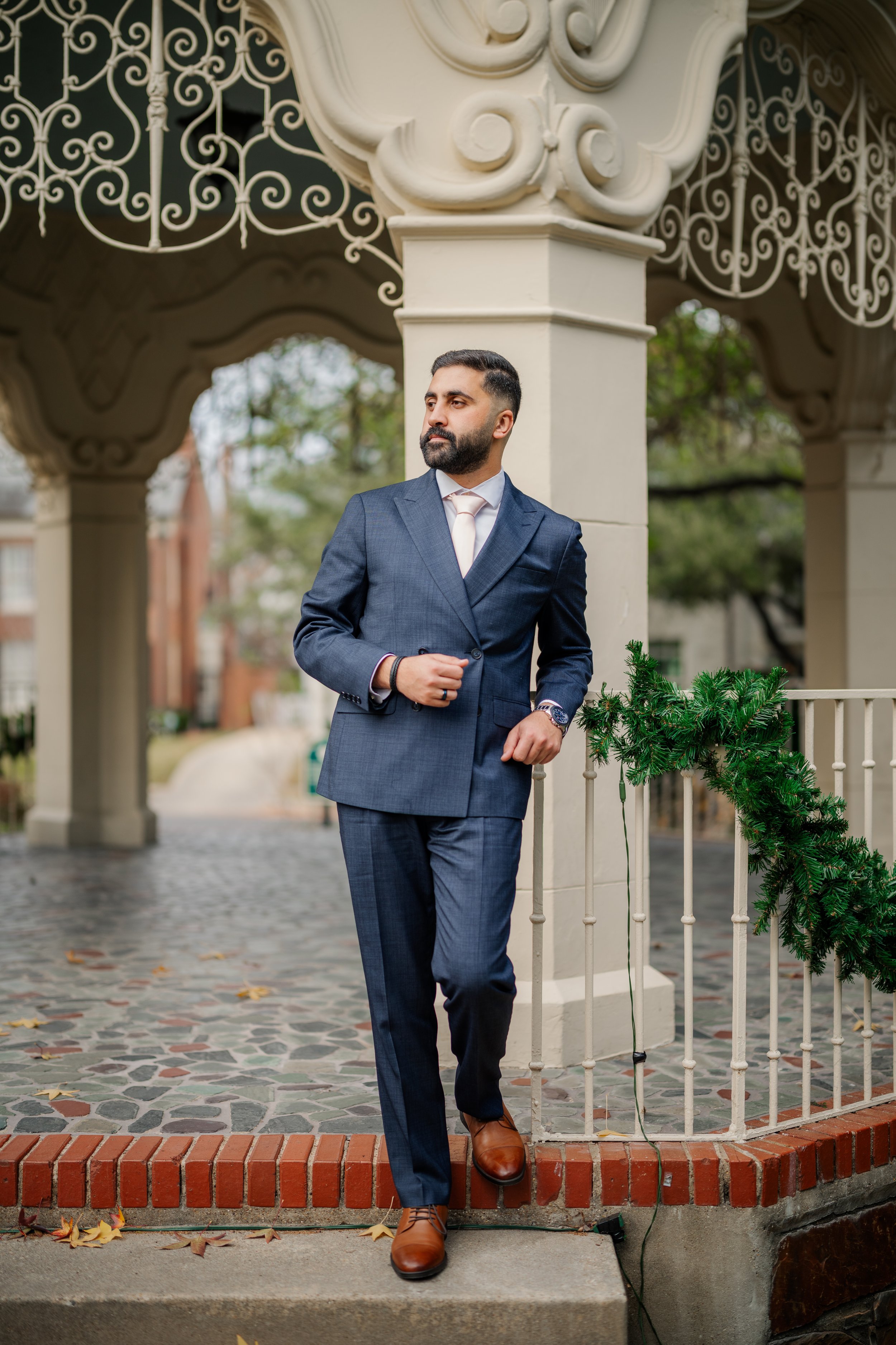 A man in a blue suit and brown shoes standing outdoors near a decorated railing with greenery, under an ornate structure with intricate ironwork.
