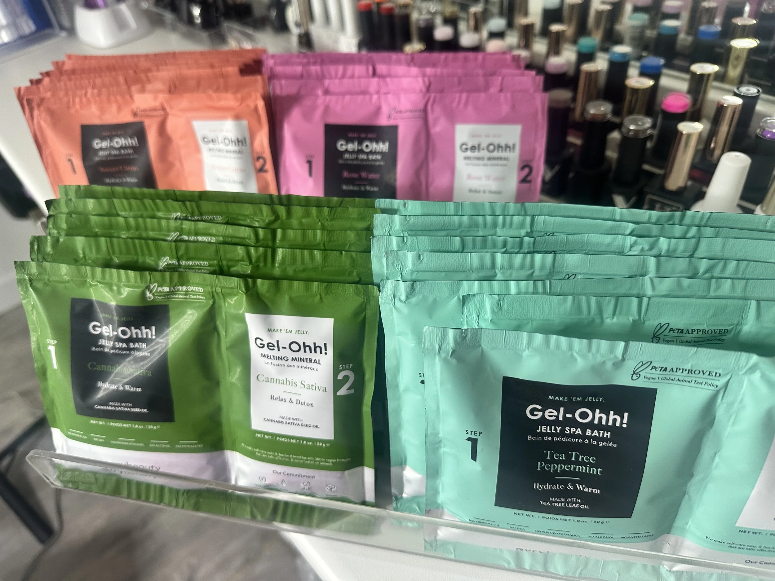 Collection of colorful packages of Gel-Ohh! Jelly Spa Bath products on display, including green, pink, and coral packets, with a background of various nail polish bottles.