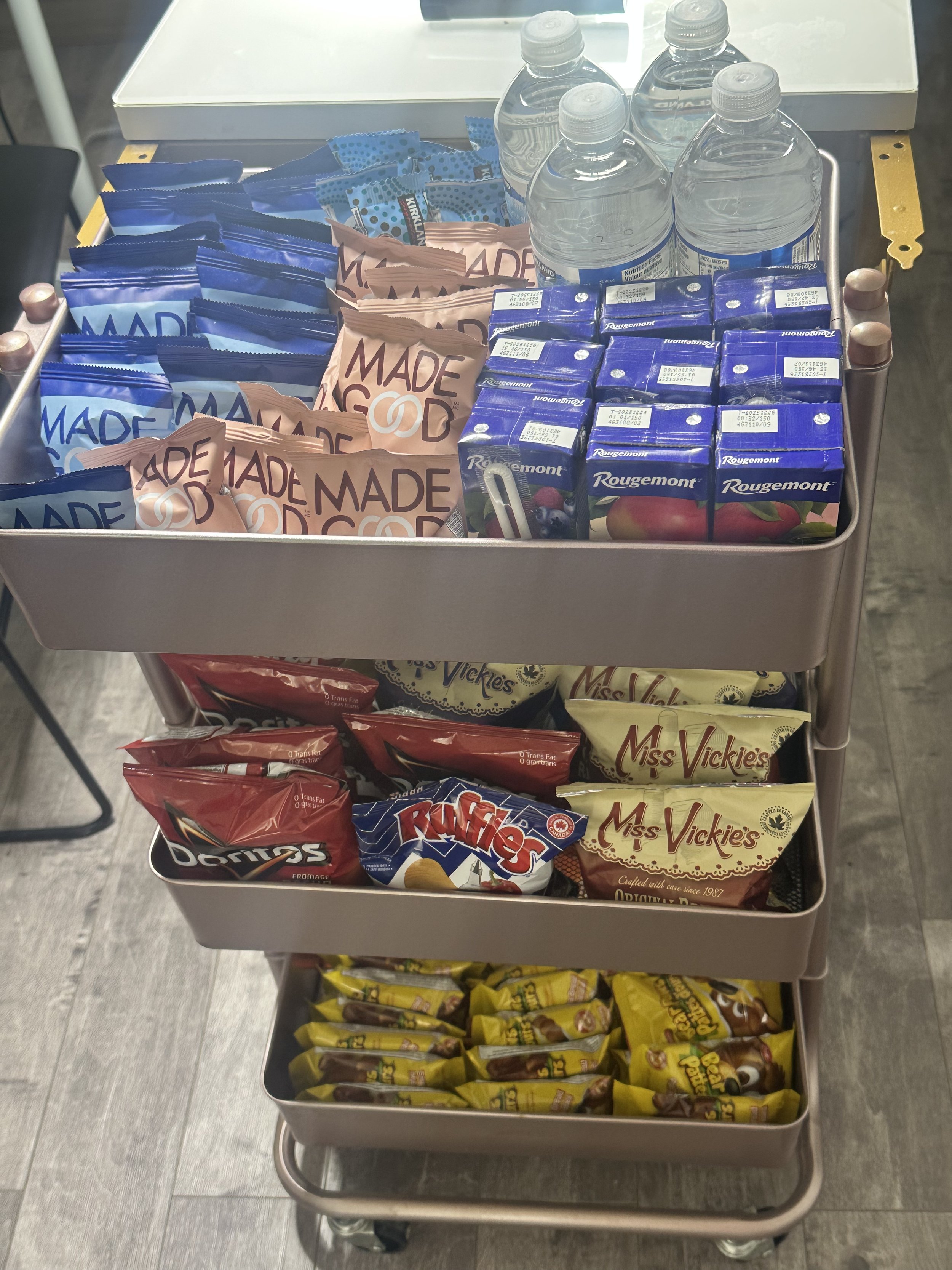 A three-tiered metal snack cart filled with bottled water, juice boxes, and various snack bags including chips, candy, and cookies.
