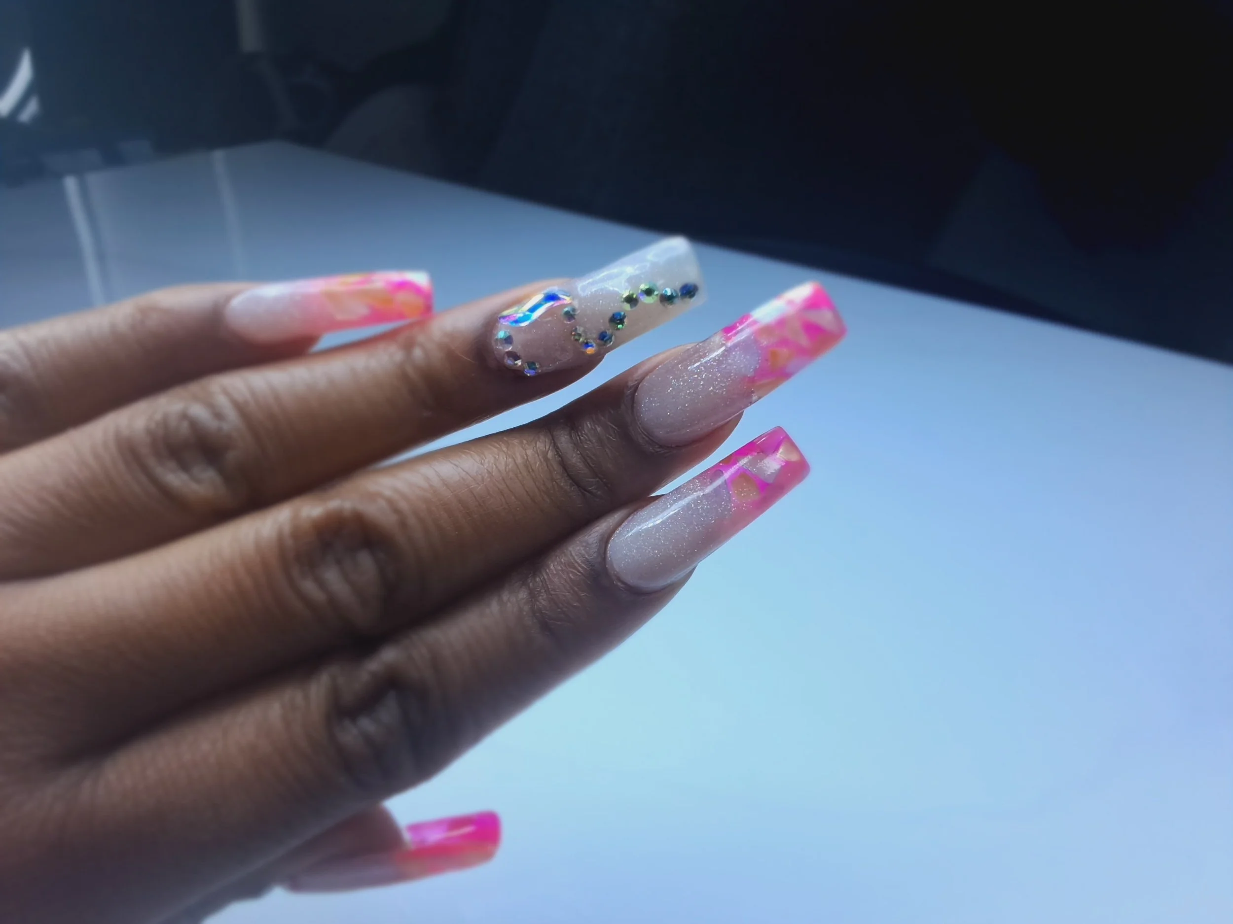 Close-up of a hand with long, decorated acrylic nails featuring pink, white, and orange marble patterns and rhinestone embellishments.