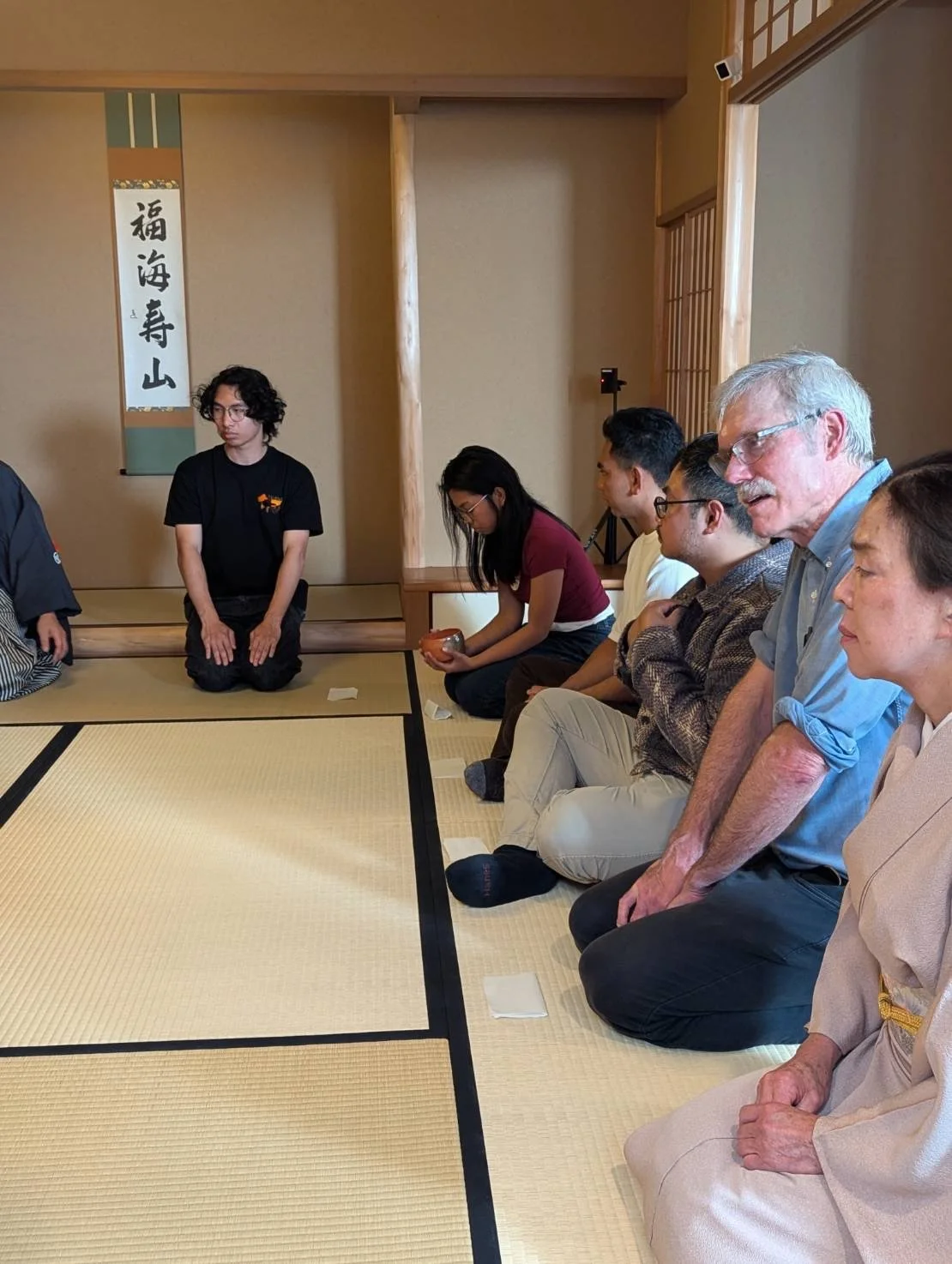Group of people sitting in a traditional Japanese tatami room, participating in a cultural activity, with a person kneeling at the front and a Japanese calligraphy scroll hanging on the wall.