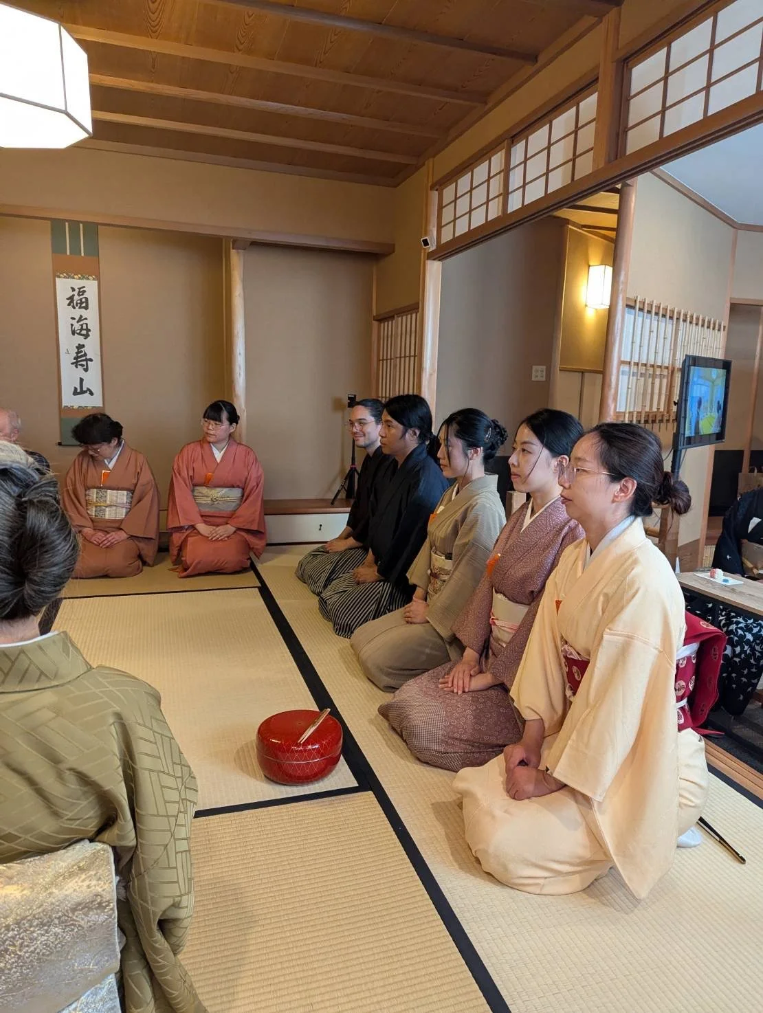 Group of people dressed in traditional Japanese kimonos sitting in seiza position during a gathering or tea ceremony inside a Japanese-style room.