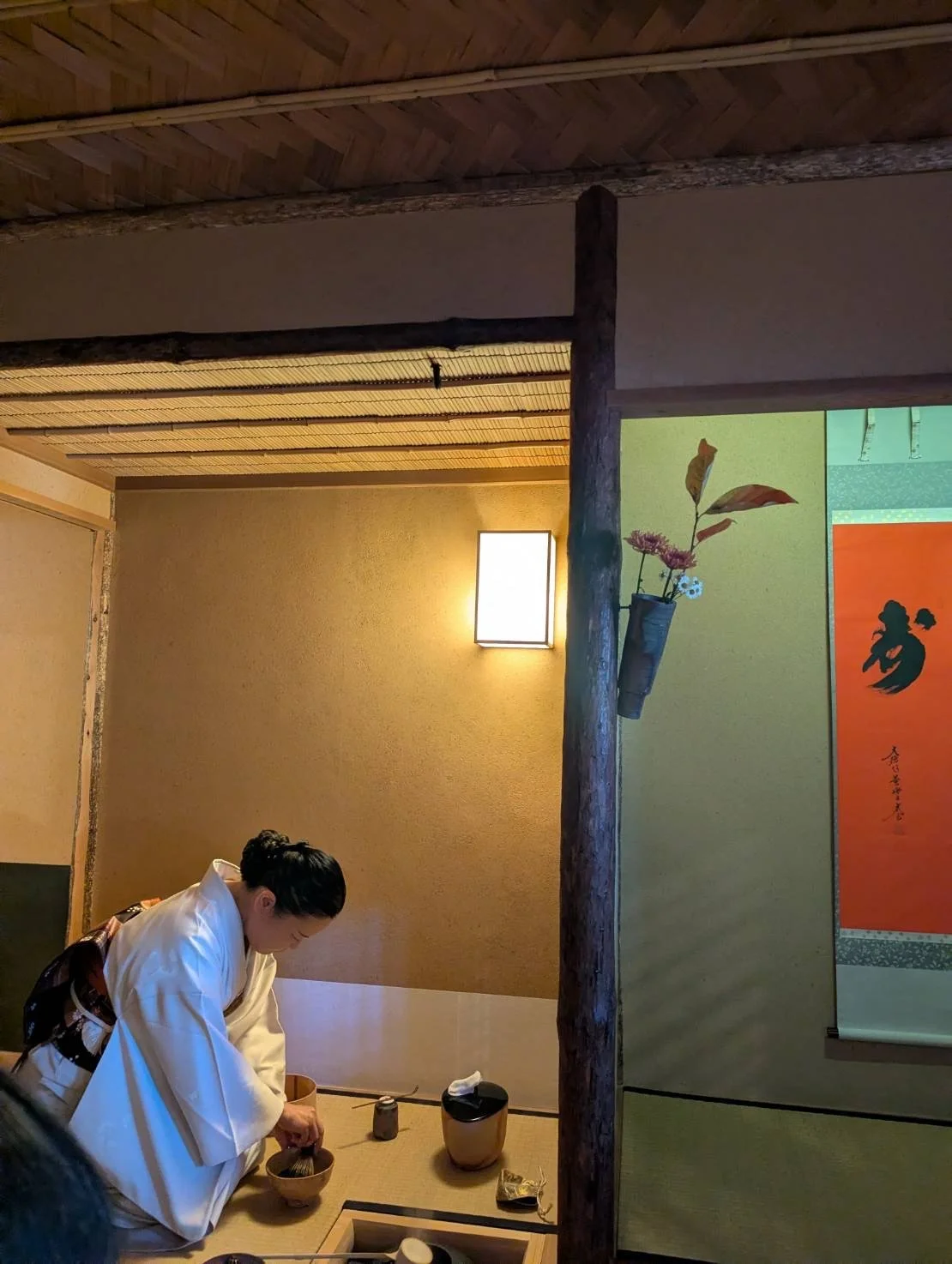 A woman dressed in traditional Japanese attire preparing tea in a tatami room with wooden beams, a wall sconce, and a hanging scroll with calligraphy, along with a vase holding flowers mounted on a wooden post.