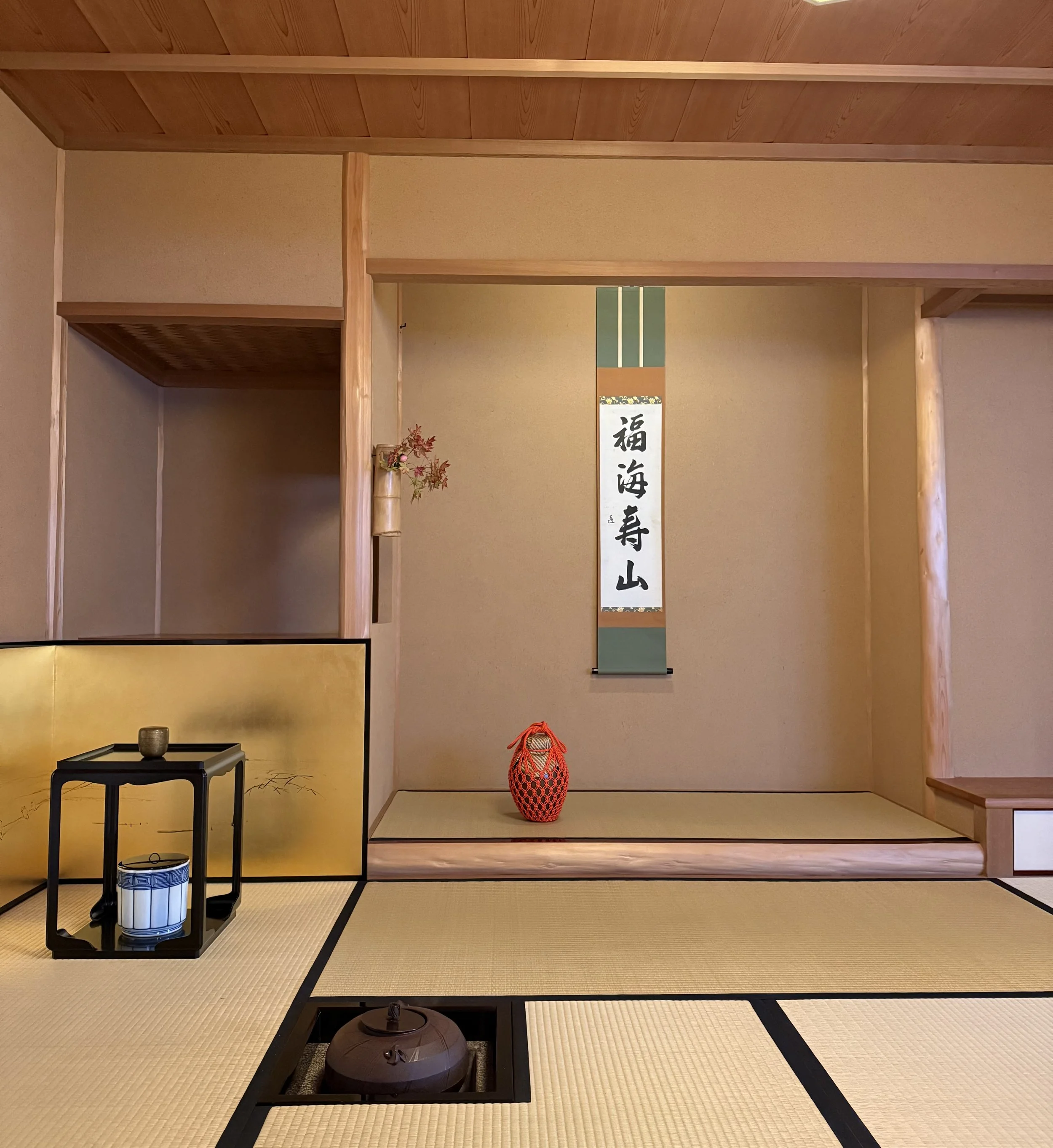 A traditional Japanese tokonoma alcove with a hanging scroll, a red woven vase, and a small flower arrangement. The room has tatami mat flooring and a gold folding screen in the background.