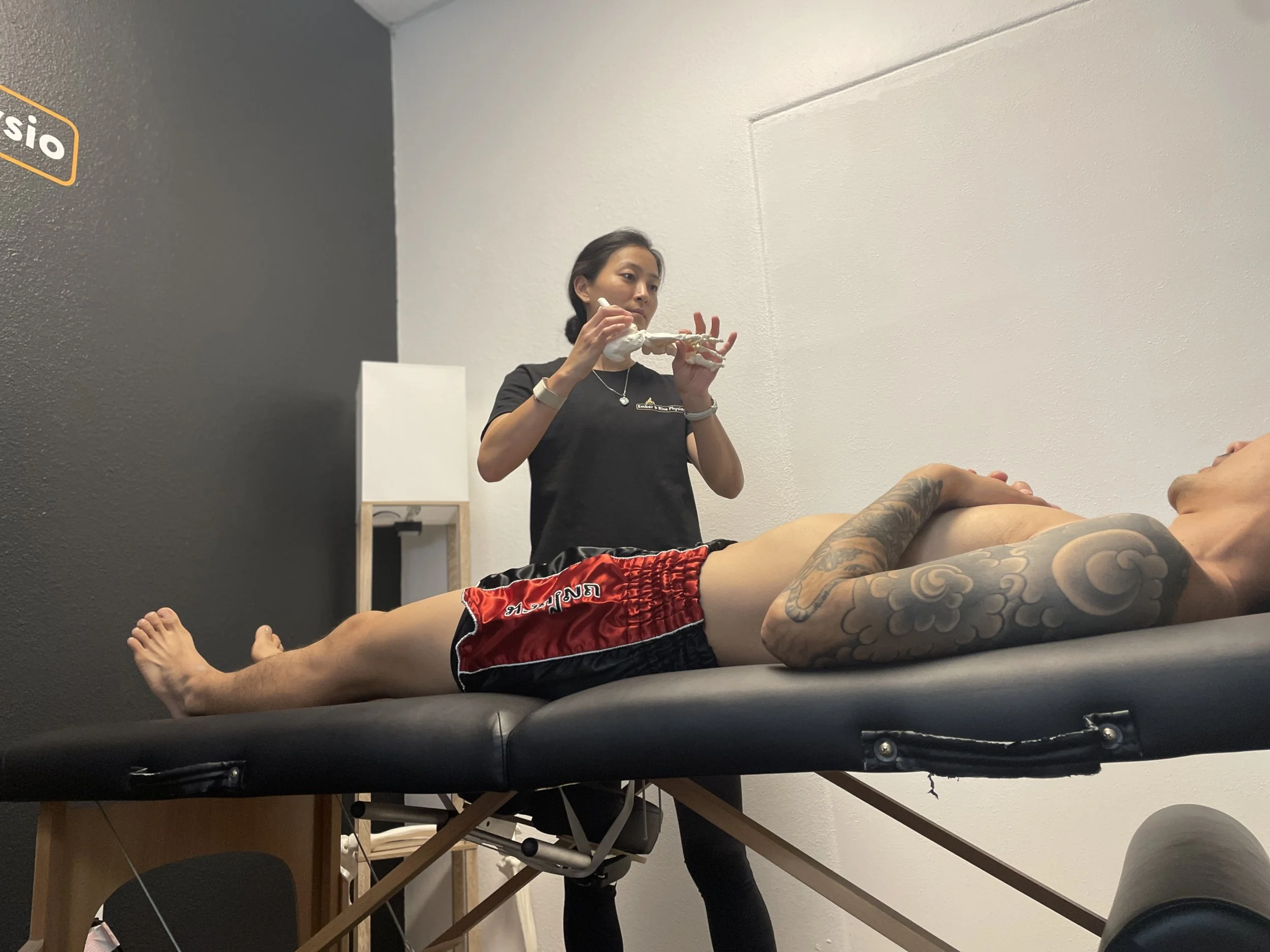 female physical therapist explaining foot and ankle biomechanics while holding a foot anatomy model to a male muay thai athlete wearing red muay thai shorts lying down on a table