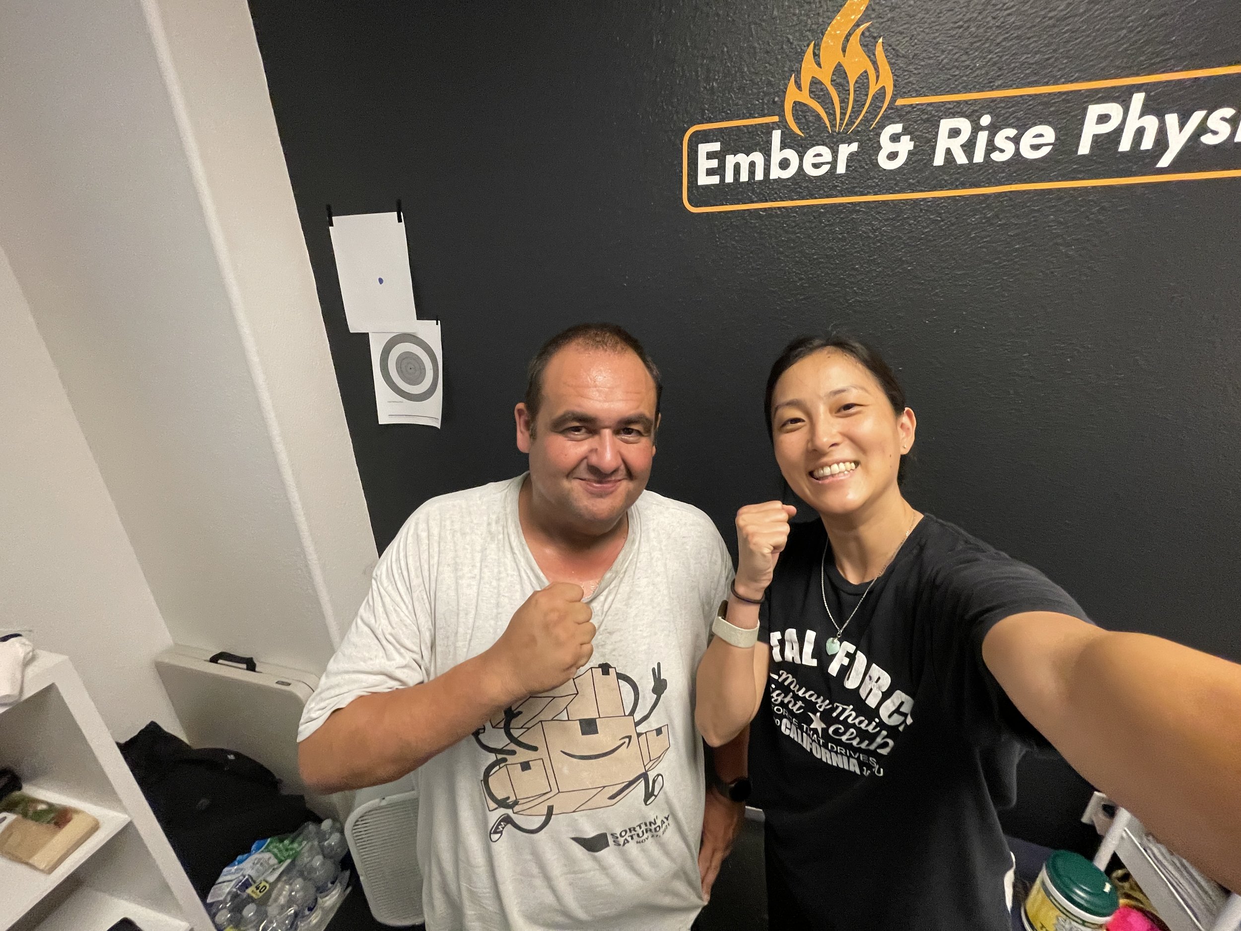 female physical therapist and client smiling at camera posing with fist wearing black muay thai shirt after PT session