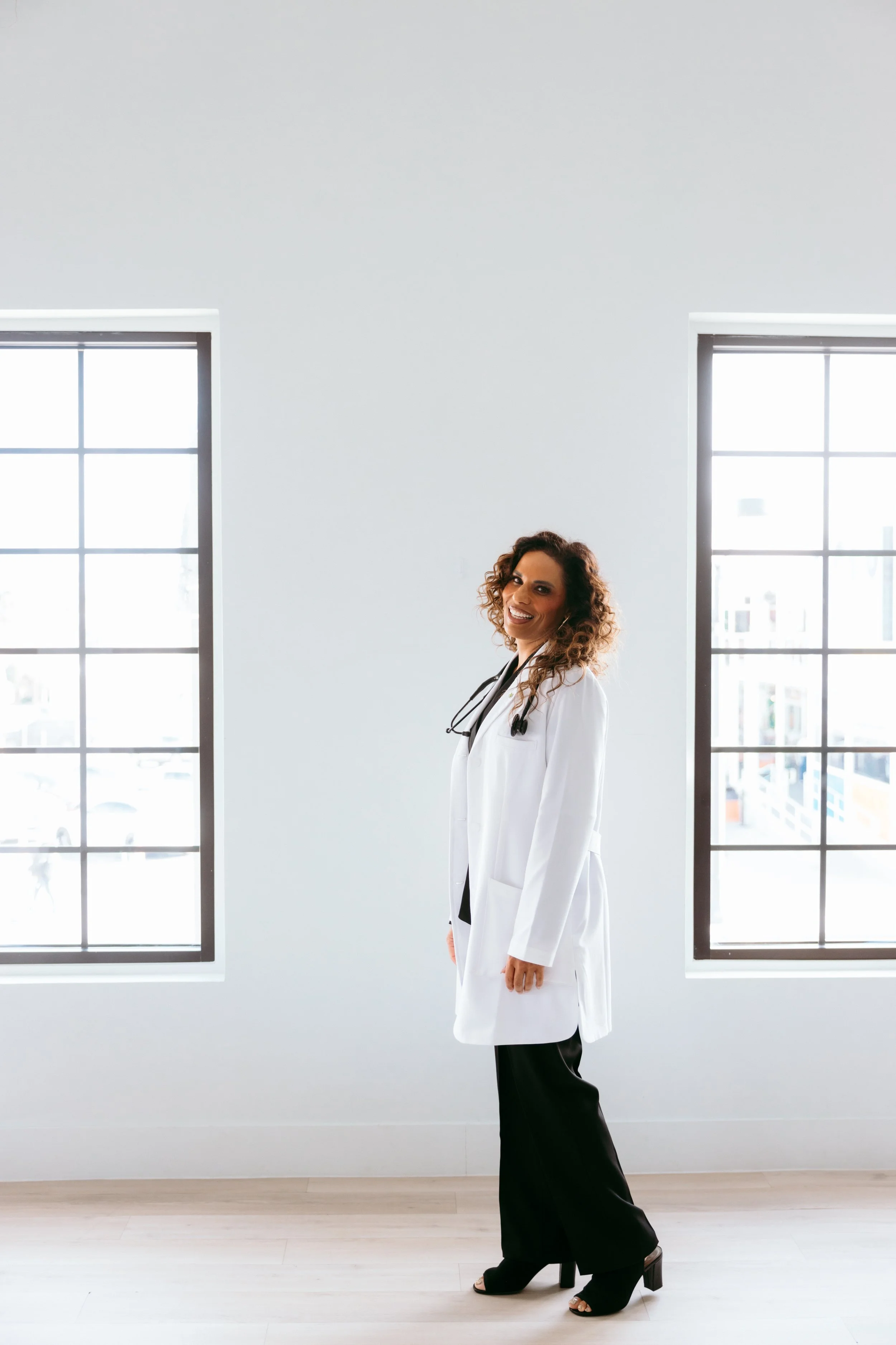 A female doctor standing in a bright room with large windows, wearing a white coat and black pants, smiling.