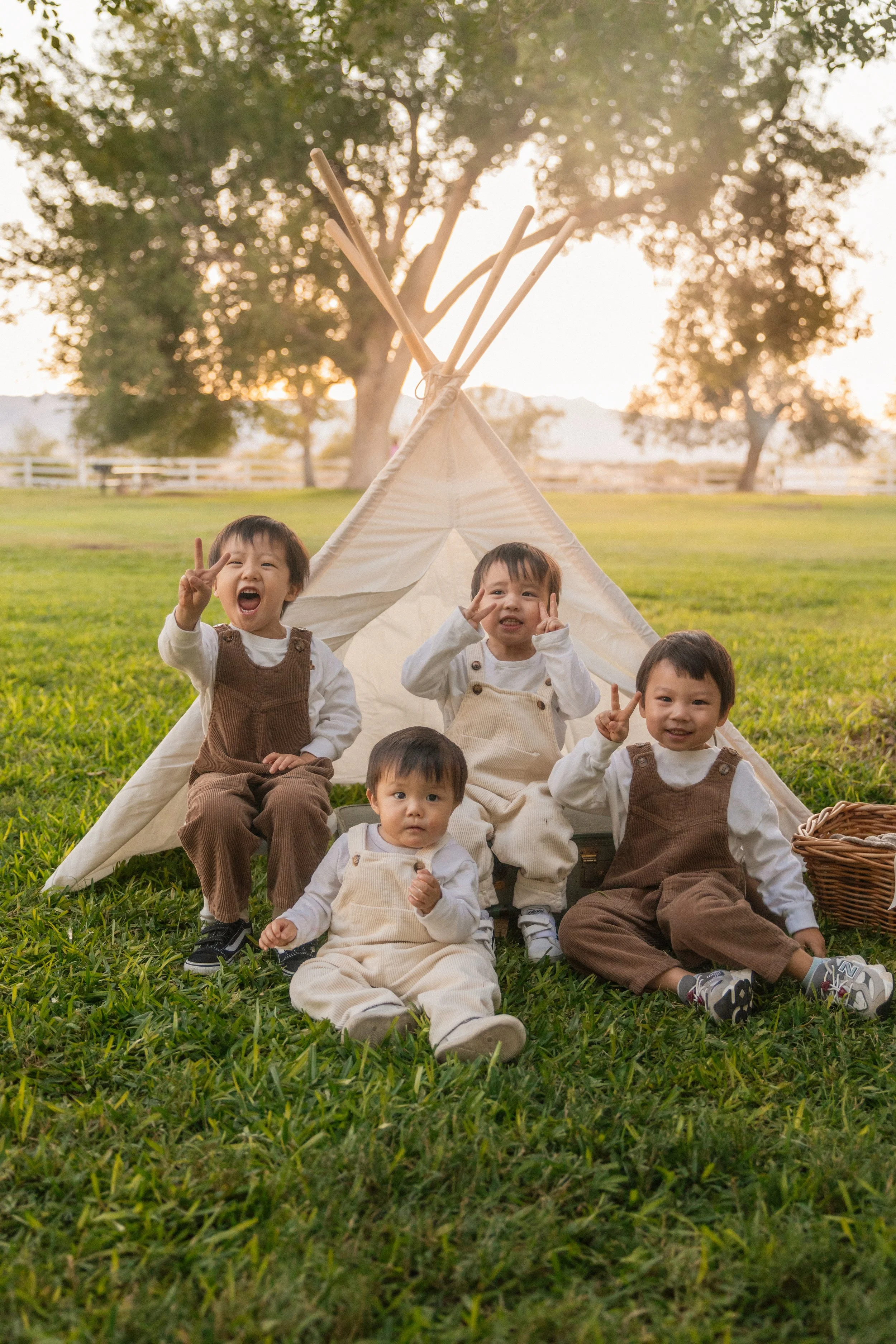 Five young children sitting in front of a small teepee tent on a grassy field during sunset, playing and smiling.