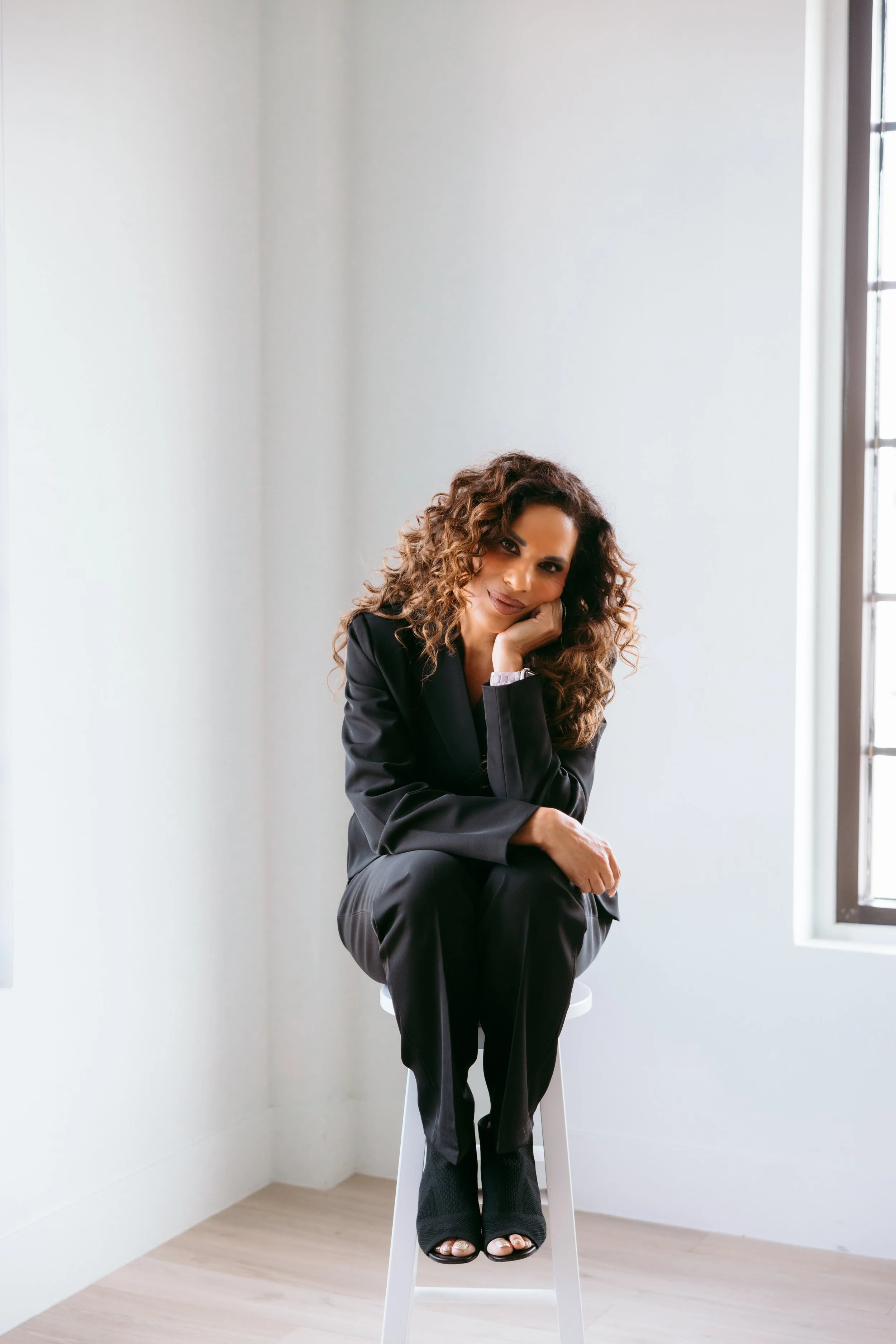 A woman with curly brown hair wearing a black suit sitting on a white stool in a bright room with a large window.