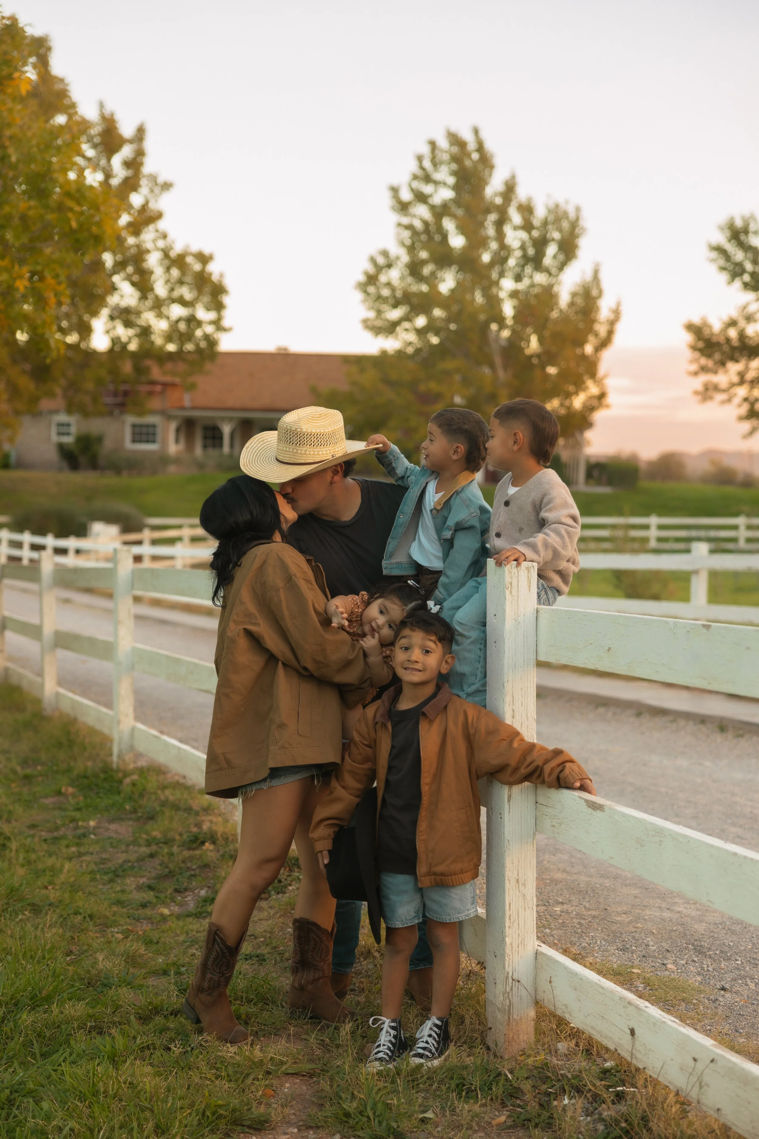 Family of six enjoying a moment outdoors near a white fence at sunset, with a house and trees in the background.