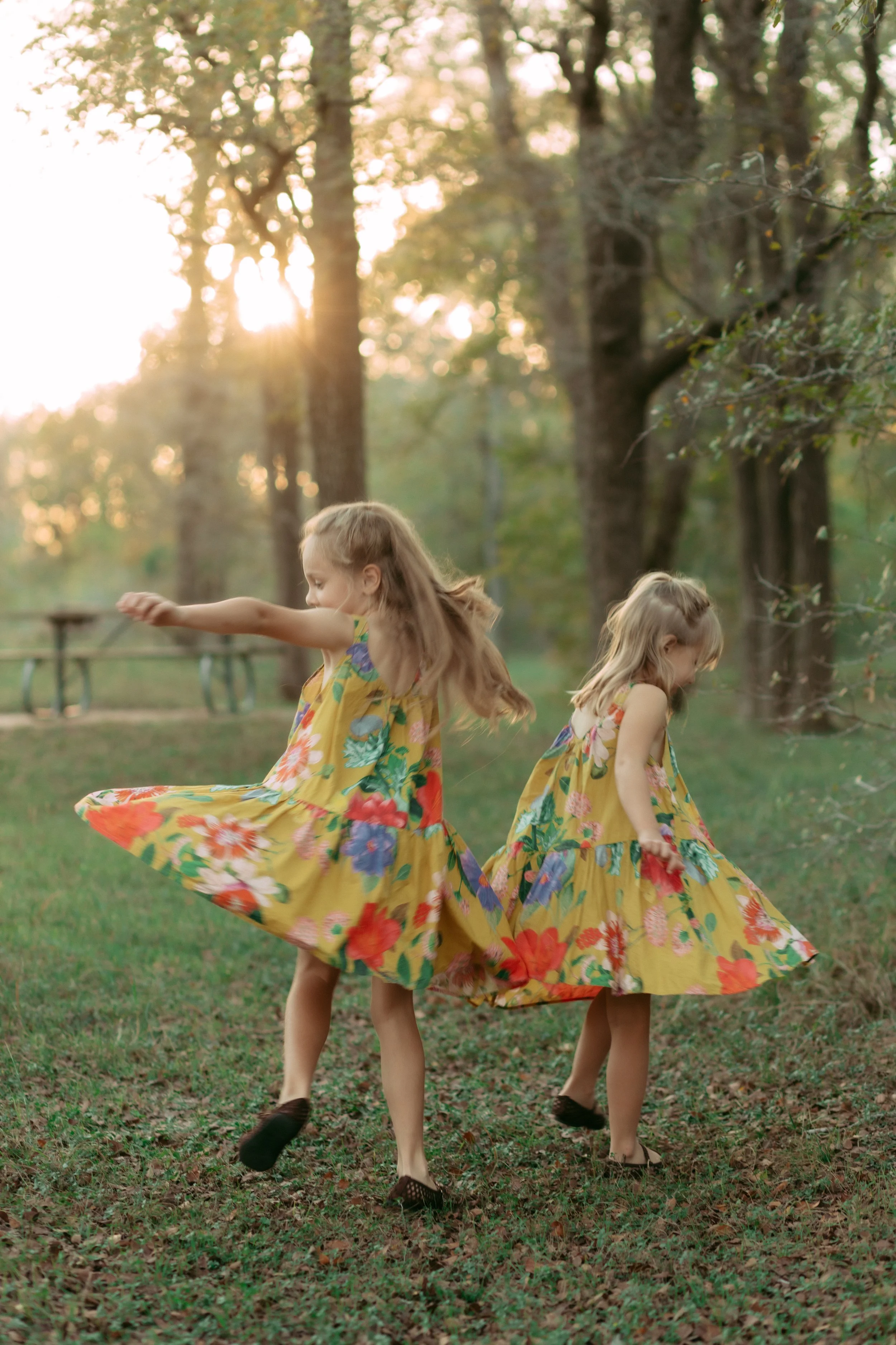 Two young girls in yellow floral dresses spinning and twirling in a wooded outdoor area during sunset.