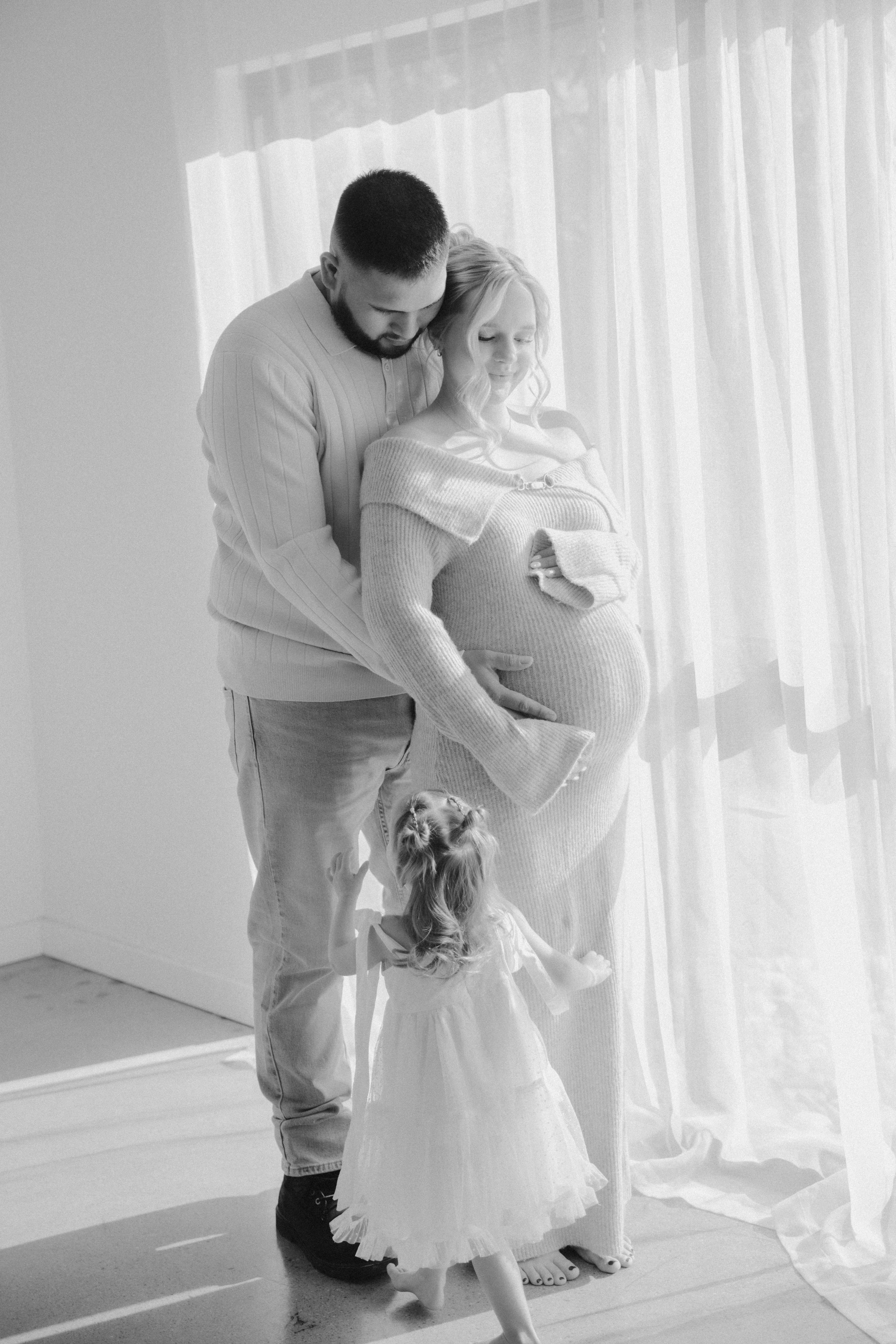 A pregnant woman and a man standing together indoors near a window with sheer curtains. The woman is holding her belly and looking down while the man is touching her belly and looking at it. A young girl in a white dress is standing in front of them,