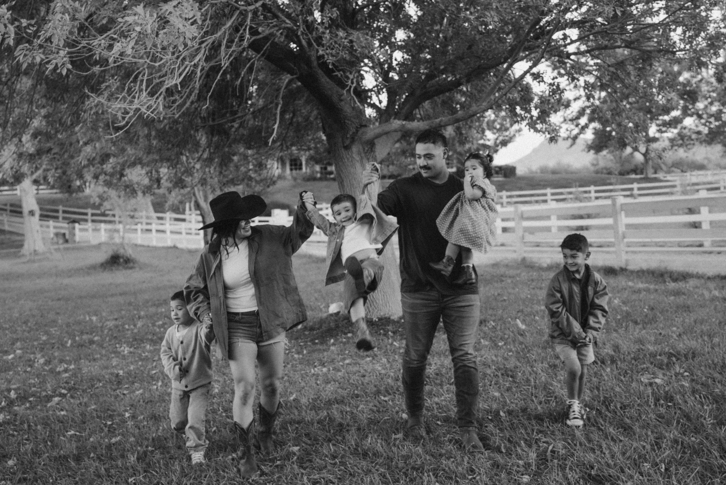 A family of five, including a woman, a man, three children, enjoying a walk outdoors in a backyard with a large tree and a white fence, in black and white.