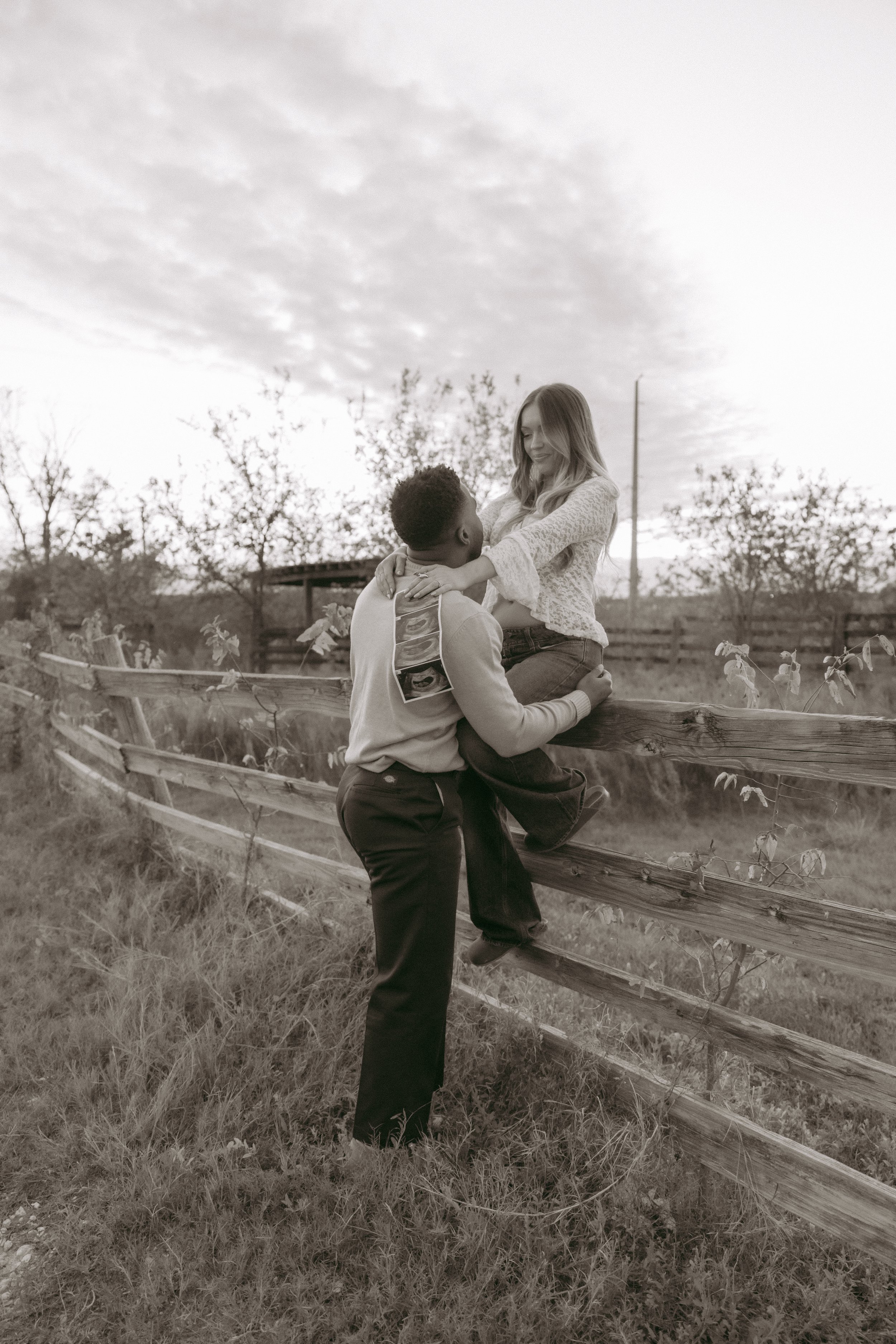 A black-and-white photo of a man lifting a woman onto a wooden fence in a rural outdoor setting with trees and a cloudy sky.