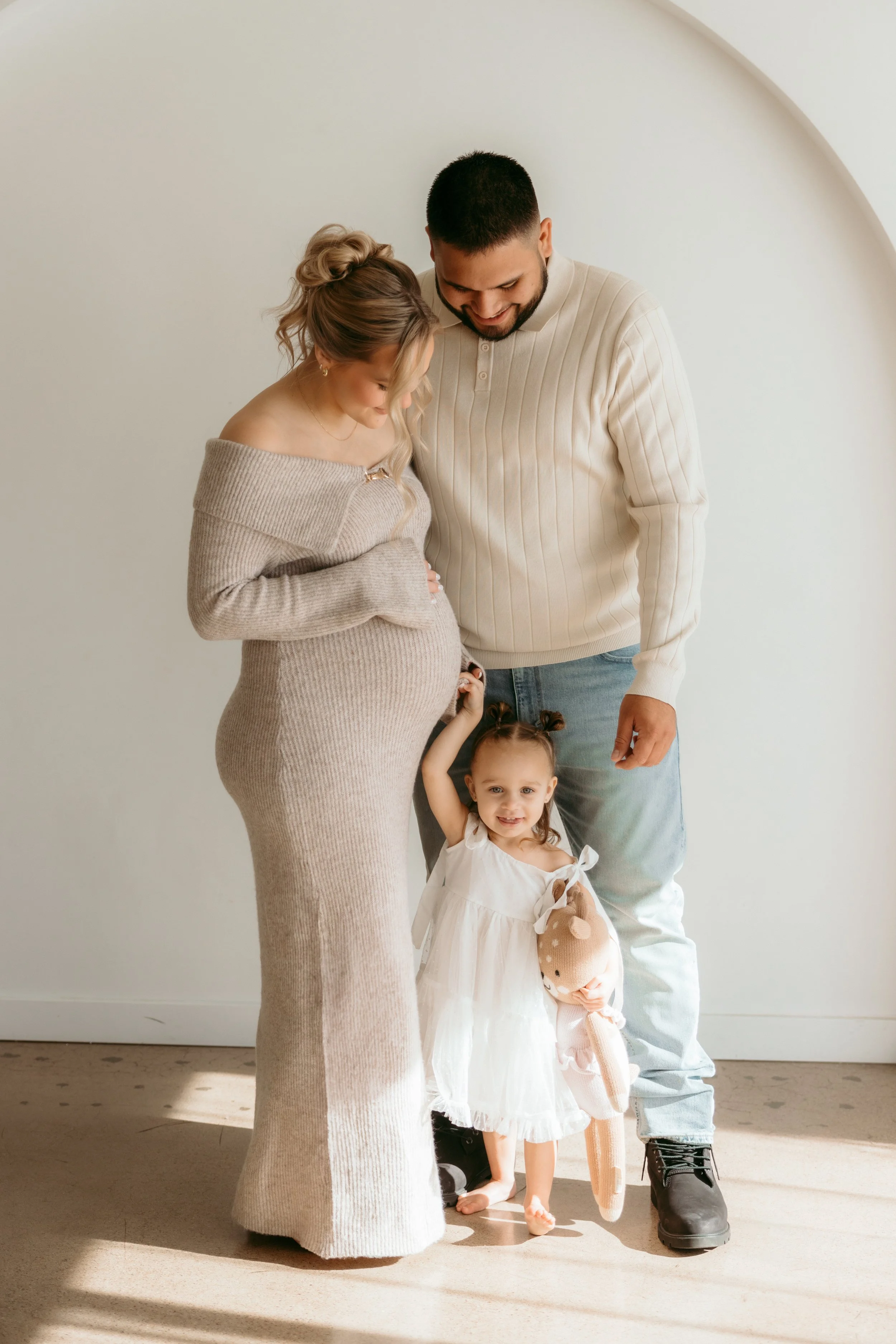 Pregnant woman and man with a young girl with pigtails standing in front of a plain white wall, smiling, with sunlight casting shadows on the floor.